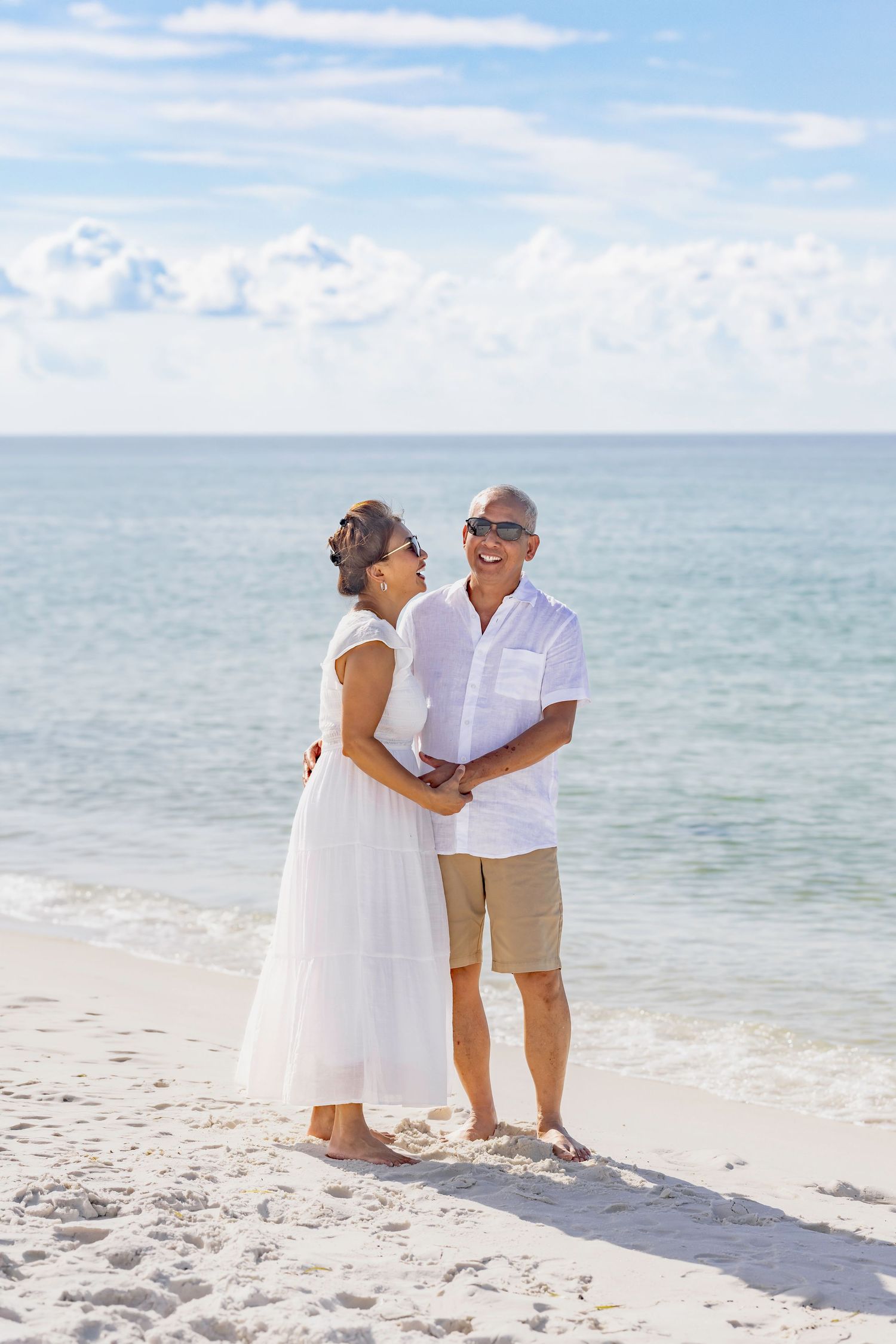 Happy mature couple holding hands on a white sand beach, dressed in white, smiling at each other with ocean backdrop.