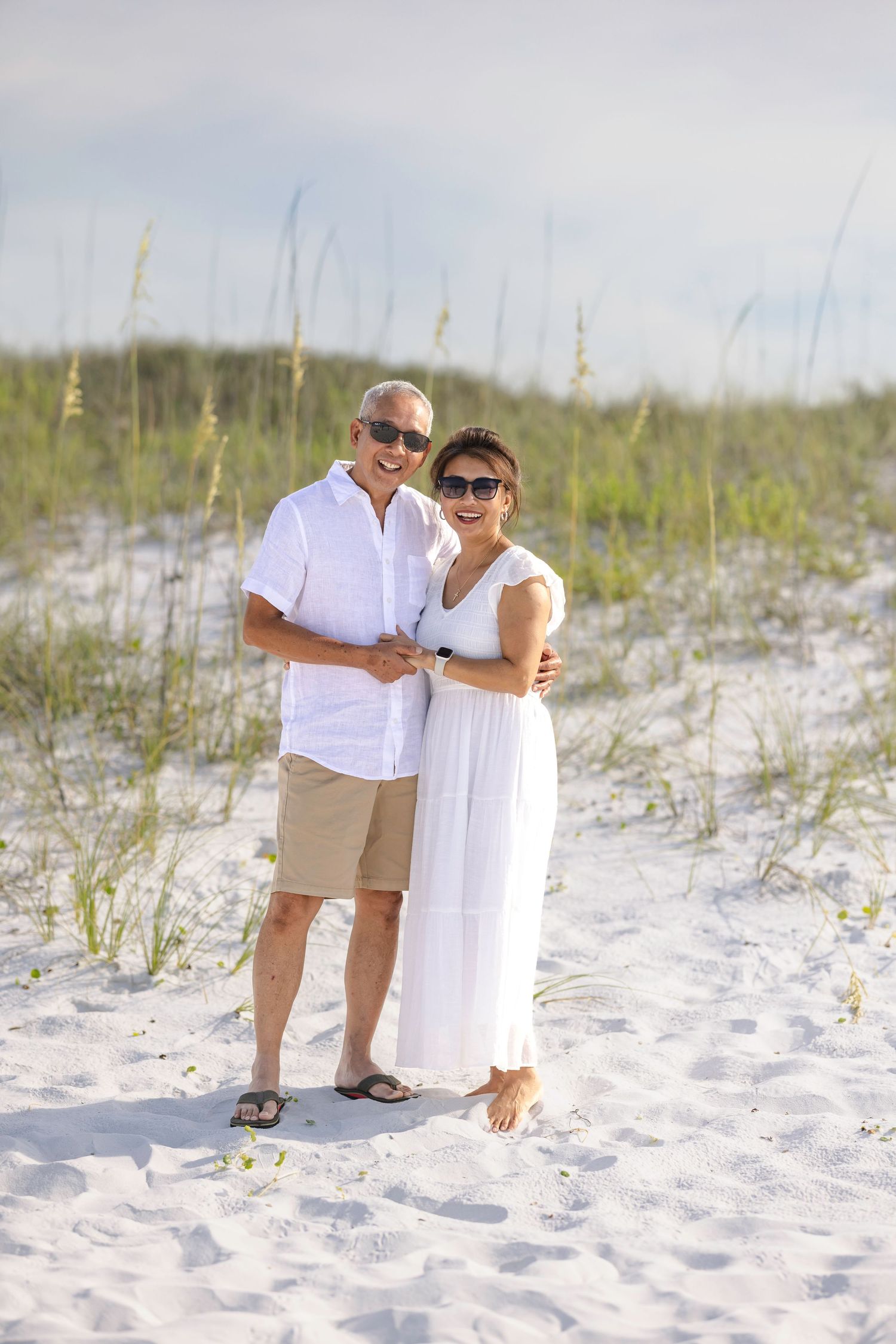 Happy mature couple in white outfits smiling together on sandy beach dunes on sunny day.