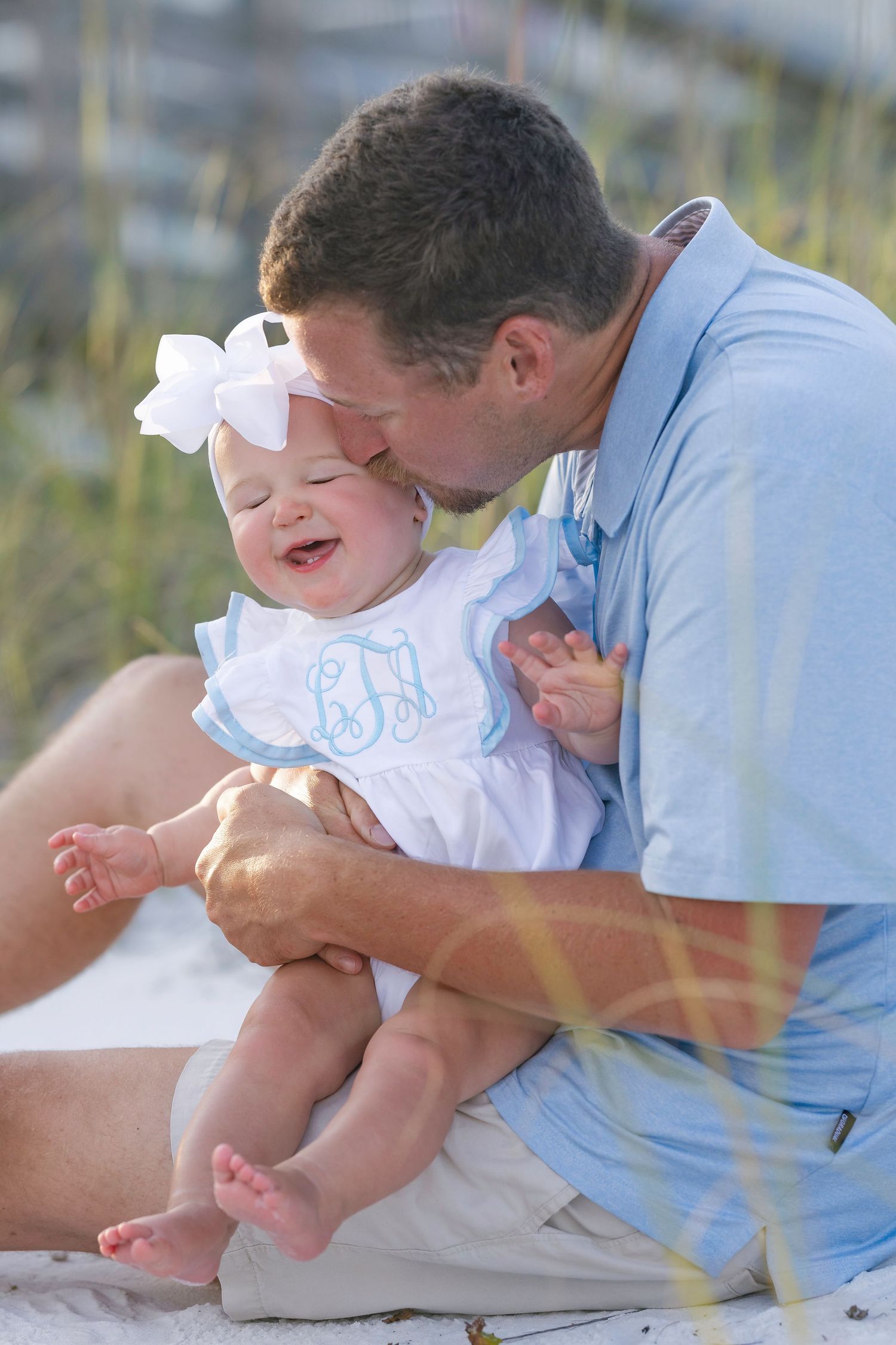 Father kissing laughing baby girl in white monogrammed dress and bow on sunny beach.
