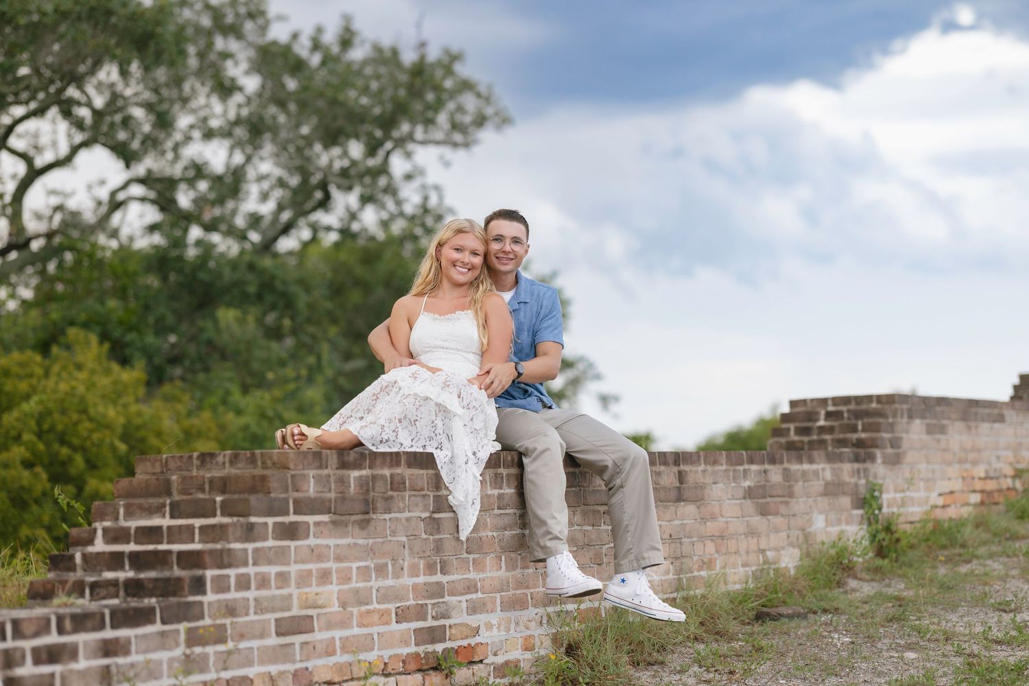 Happy couple sitting together on a rustic brick wall outdoors with lush green trees and cloudy sky behind them.