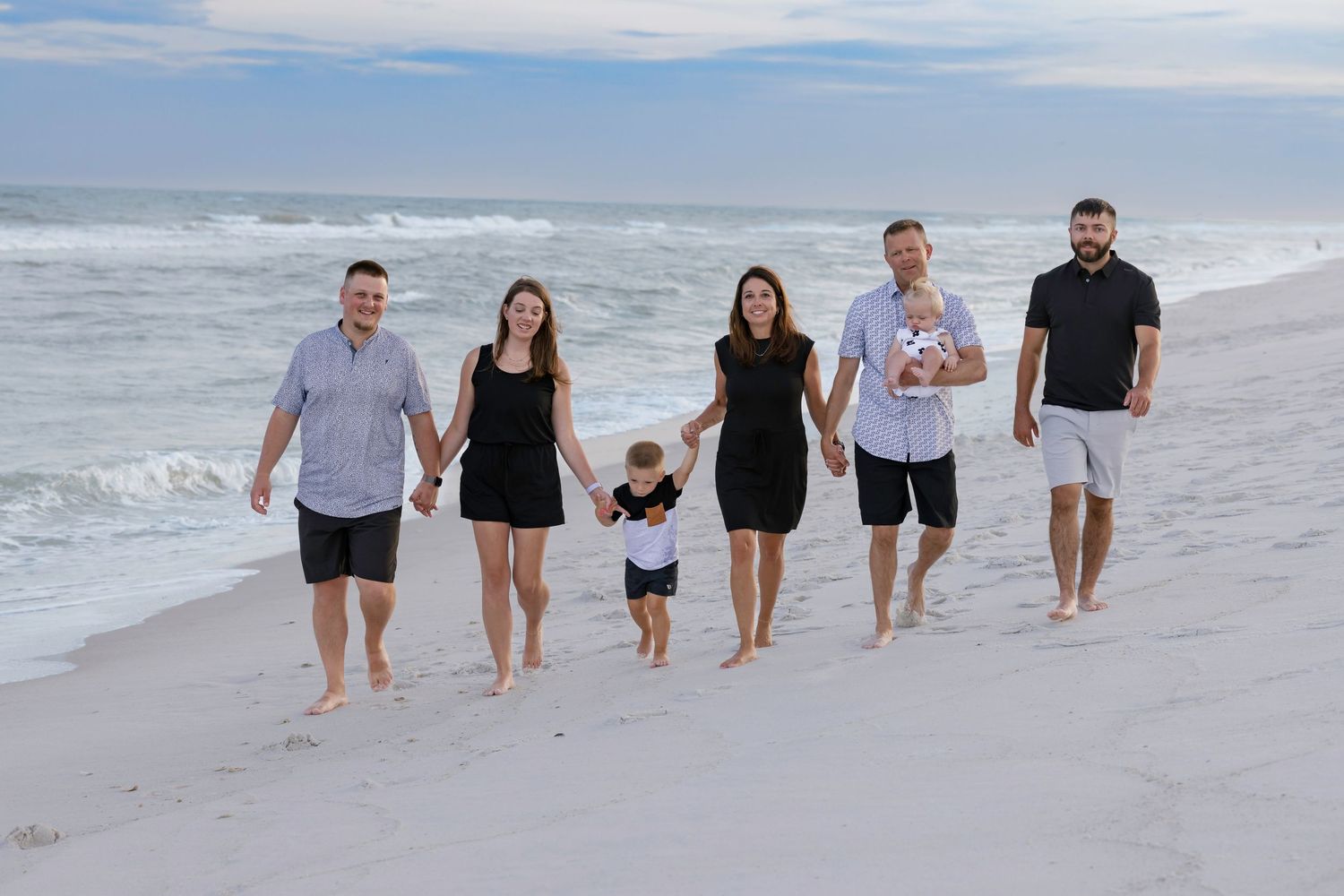 Family of six walking hand-in-hand on a sandy beach with ocean waves in the background.