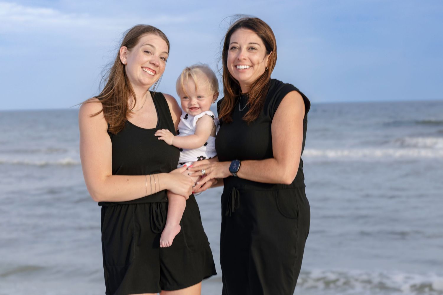 Two women in black dresses smile at the beach while holding a baby in white, with ocean waves in the background.