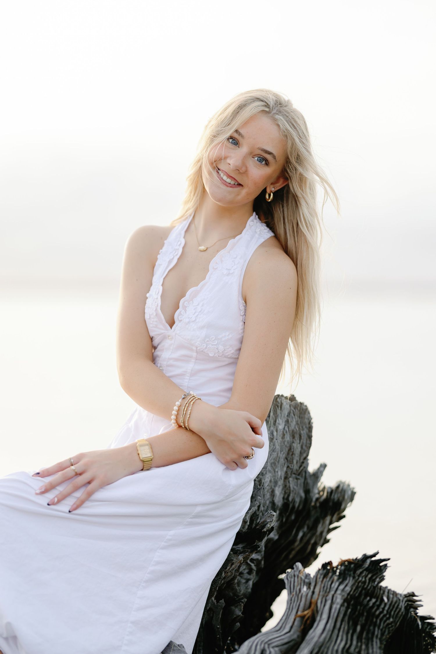 Smiling blonde woman in white lace dress sitting on driftwood at beach.