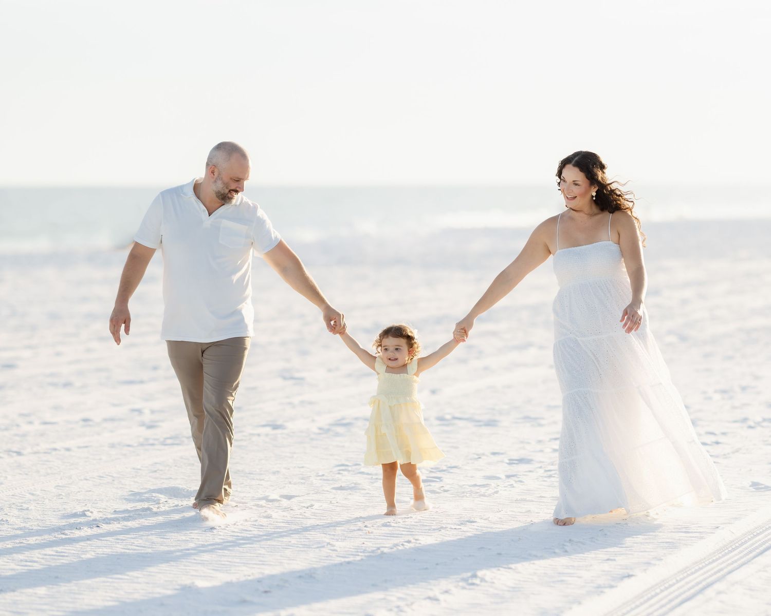 Family of three walking on white sand beach, parents holding toddler girl's hands in wedding attire.