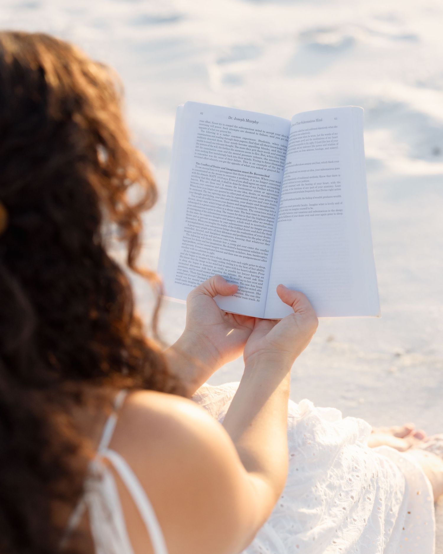 Woman with curly hair reading book while sitting on white sand beach in sunlight.