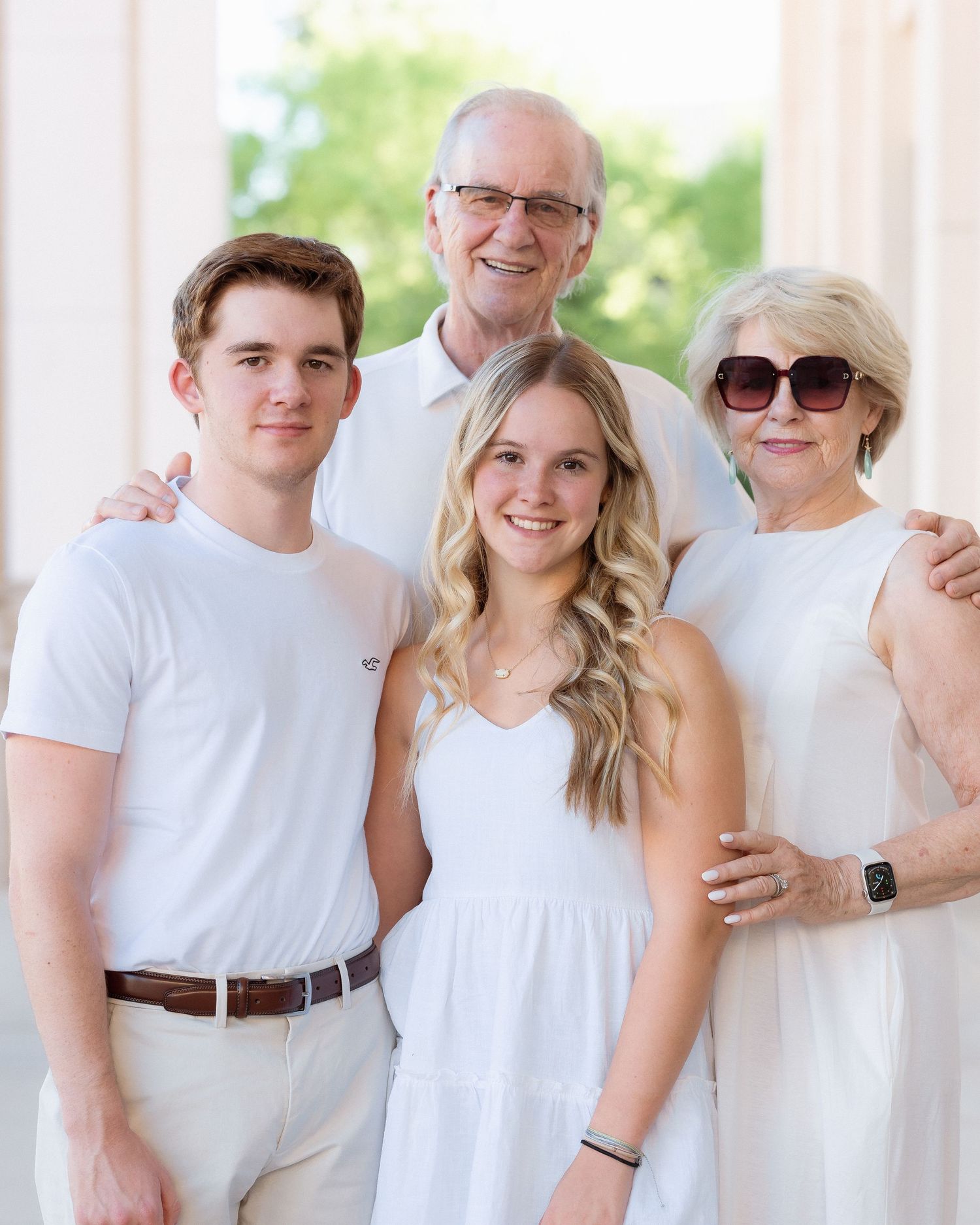 Family of four in matching white outfits posing outdoors near white columns for a formal portrait photo.