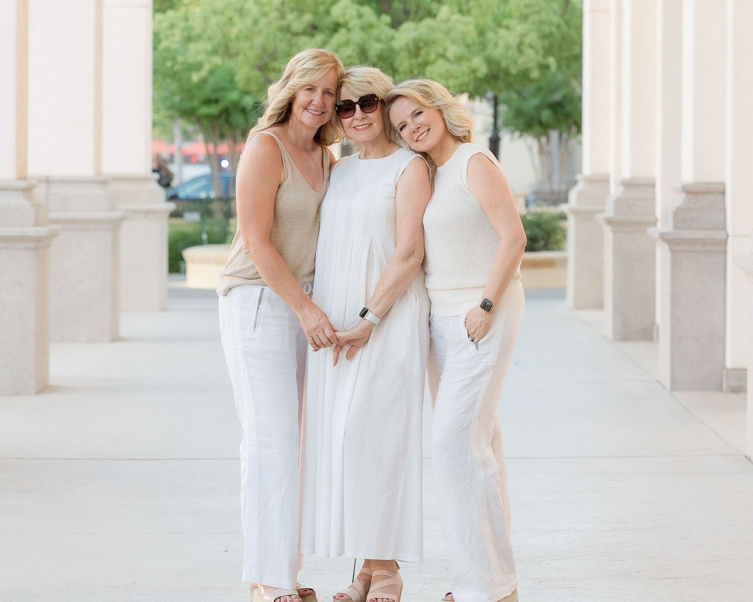 Three women in white and neutral outfits share a warm embrace during an outdoor portrait.