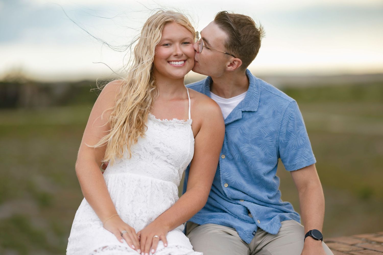 A young couple sharing a romantic kiss on a beach, woman in white dress with flowing blonde hair smiling.