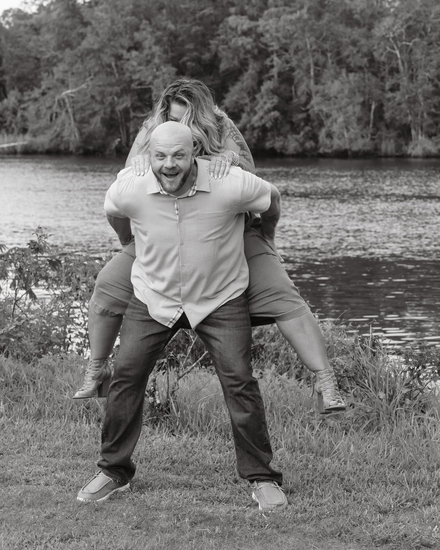 Black and white photo of a man giving a laughing woman a piggyback ride by a river.
