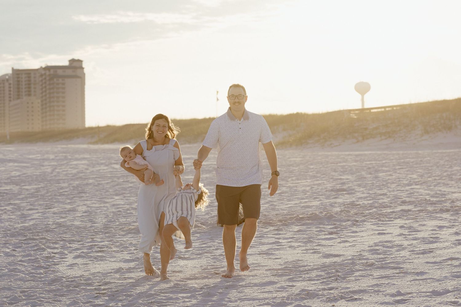 Family walking on sandy beach at sunset, parents carrying children, with coastal buildings in the background.