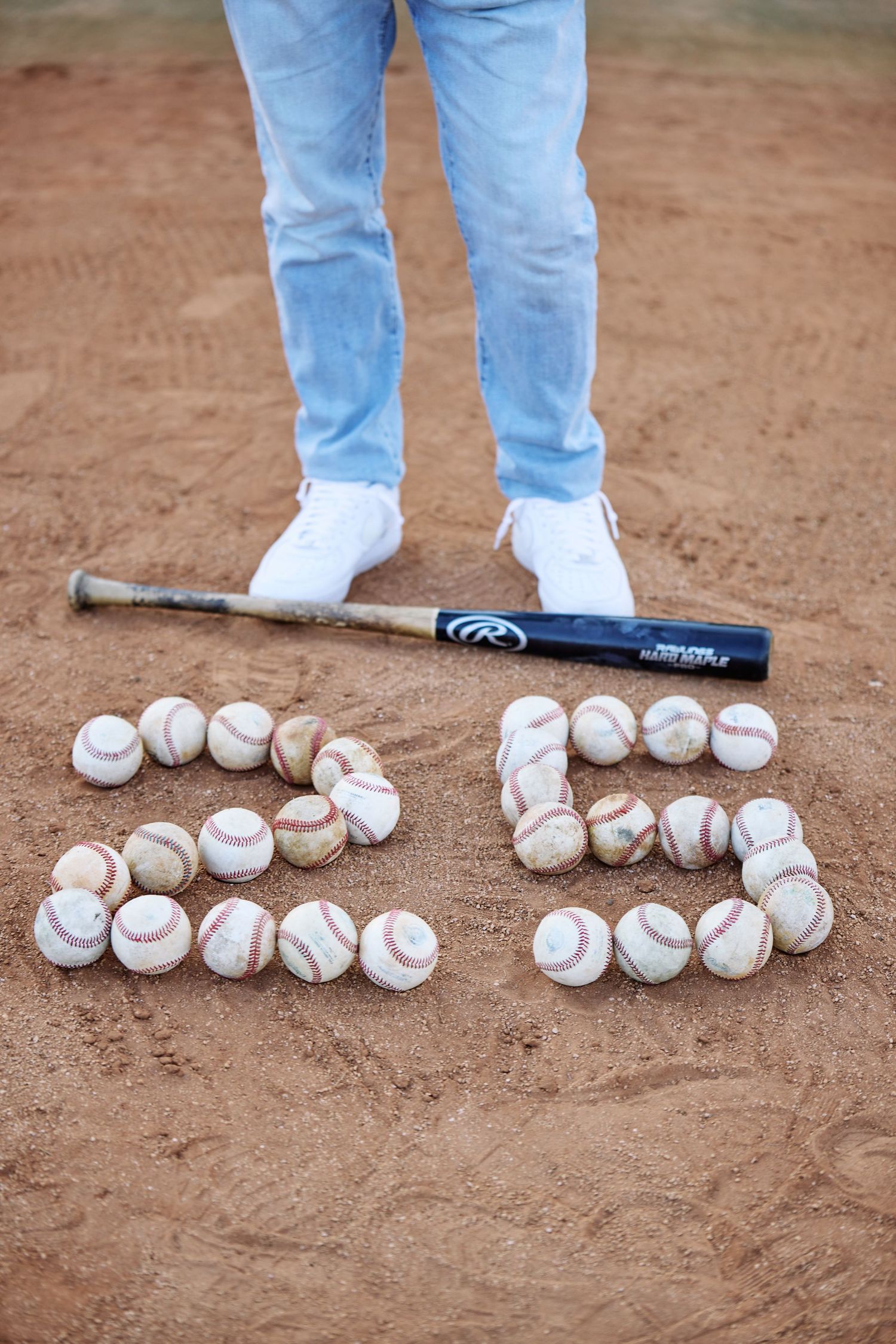 Baseball bat and baseballs arranged to form number 25 on dirt field.