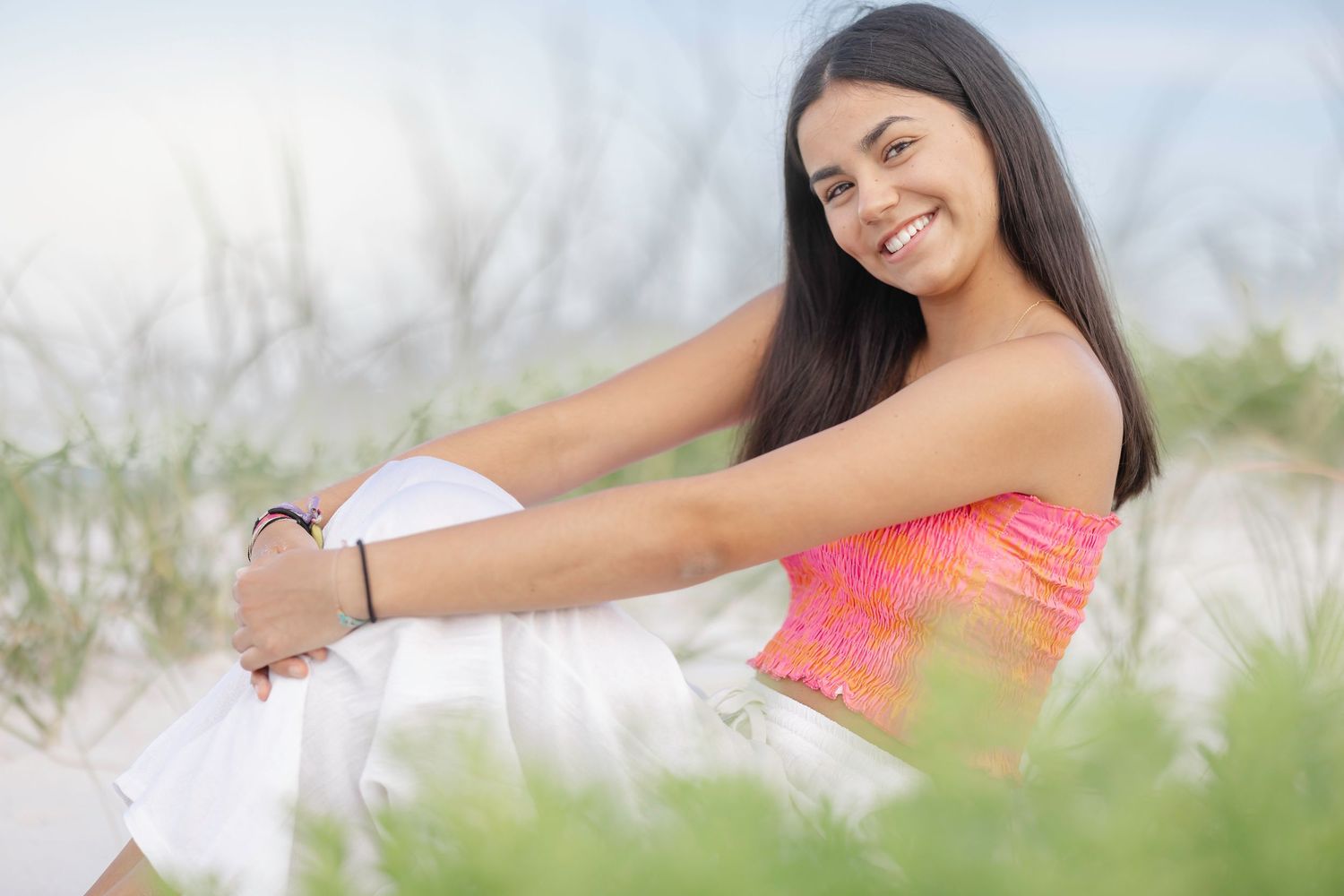Woman in bright coral top lounges on white sand beach with ocean backdrop.