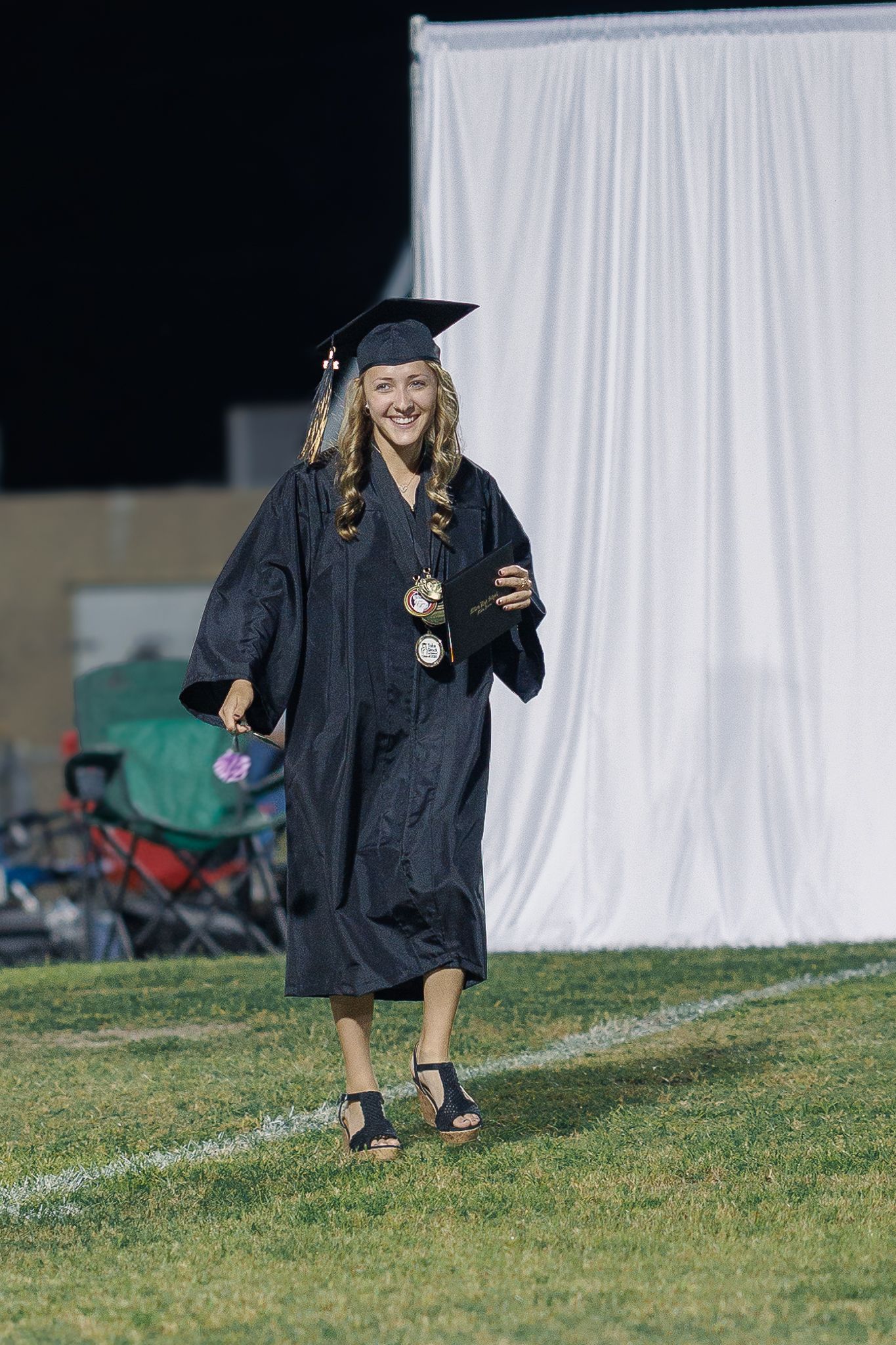 Graduate wearing black cap and gown walks across field with diploma in hand during outdoor ceremony.