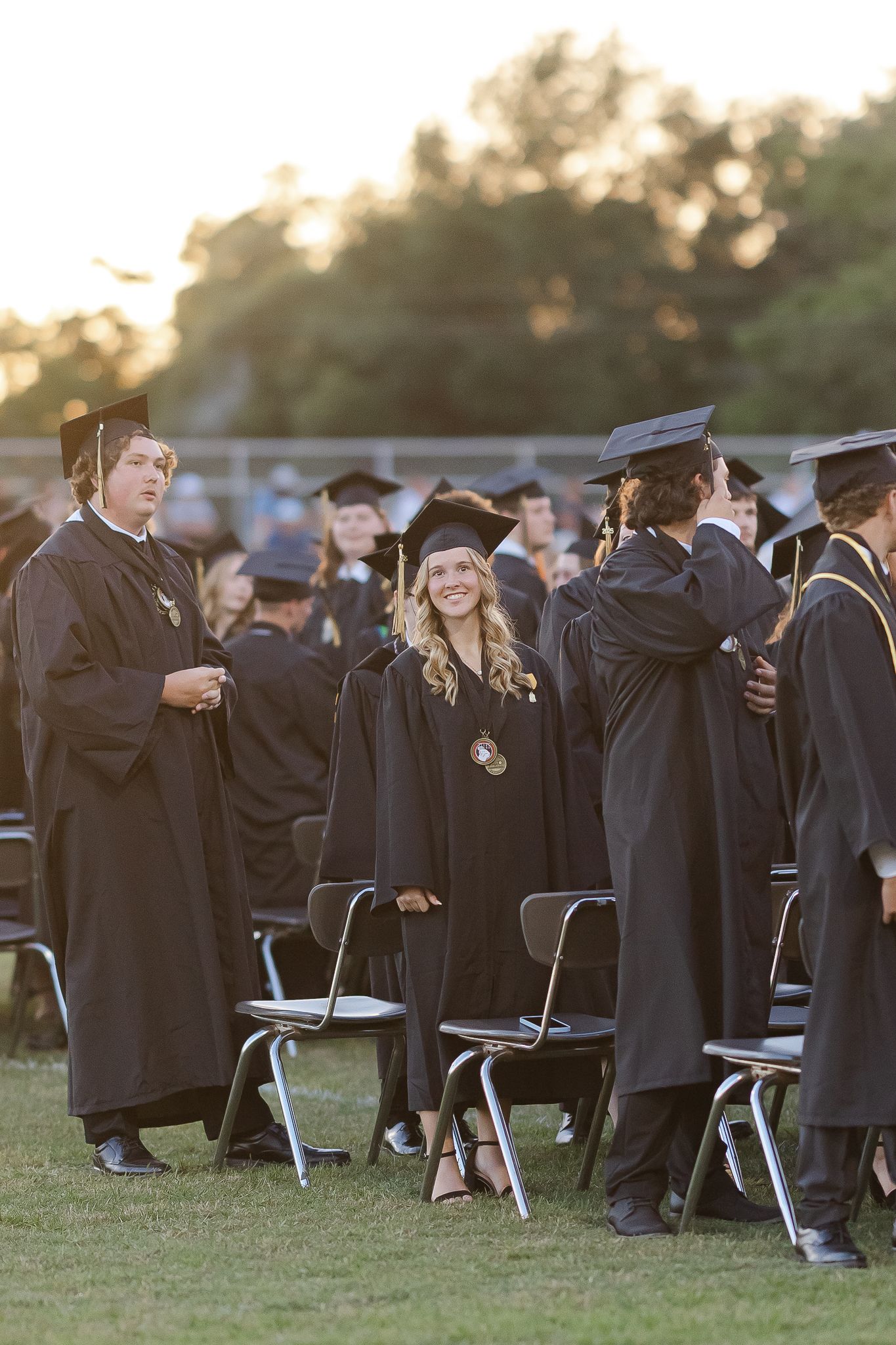 Graduates in black caps and gowns stand among folding chairs during an outdoor commencement ceremony at sunset.