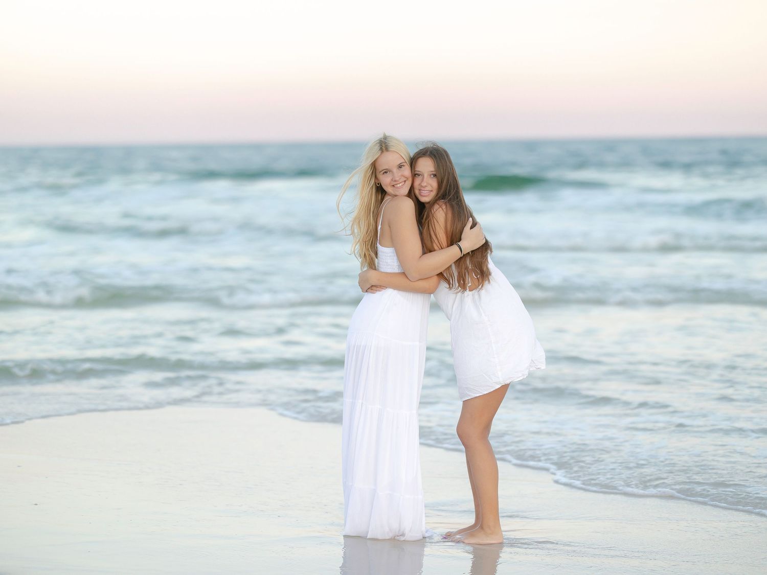 Two young women in white dresses embrace while standing at the ocean's edge during sunset.