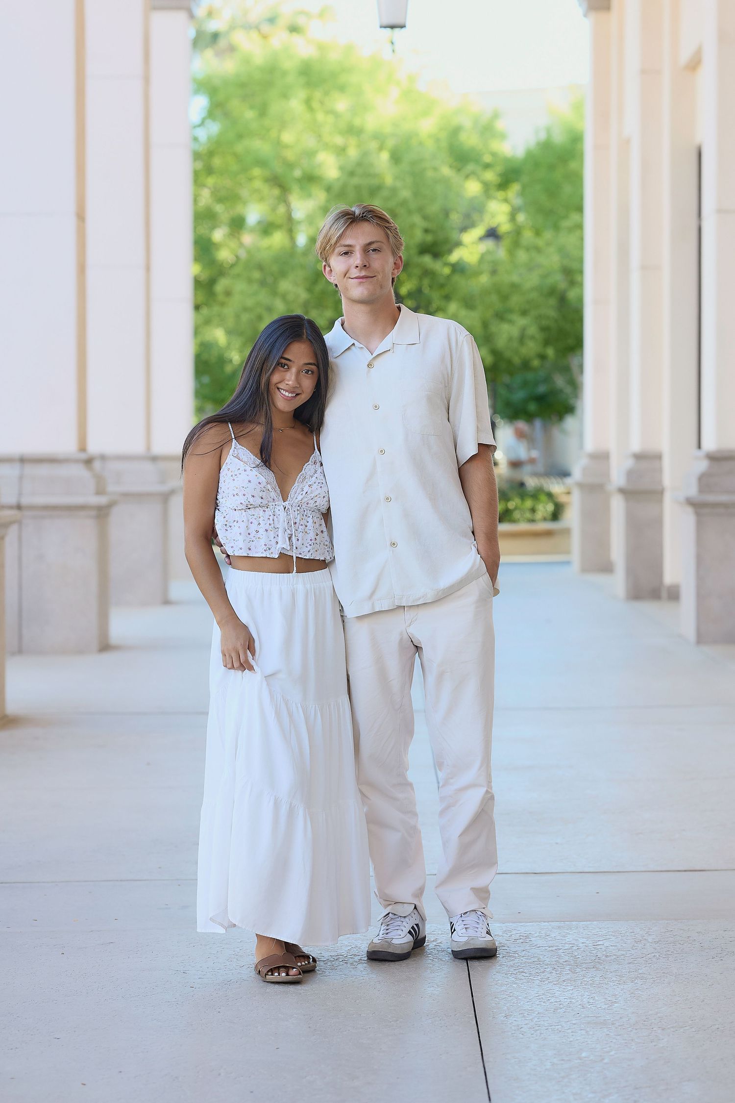 A couple wearing coordinated white outfits poses together in an outdoor colonnade setting for engagement photos.