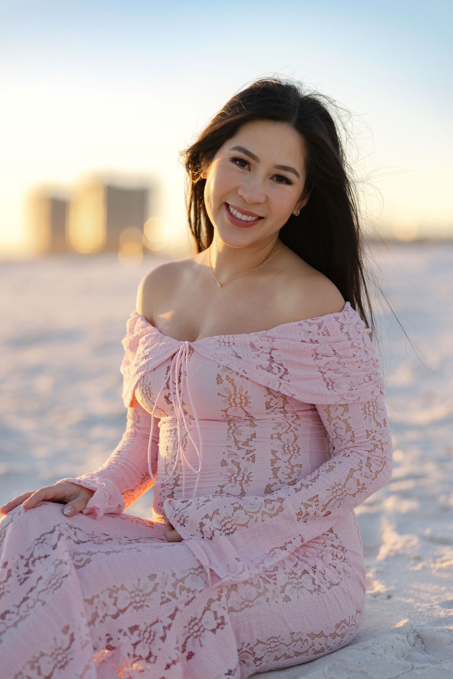 A woman in a pink lace dress poses gracefully on white sand during sunset.