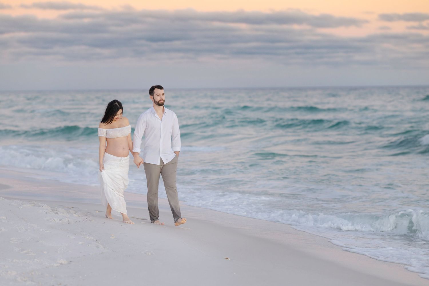 A couple walks along a sandy beach at sunset with ocean waves and soft clouds in the background.