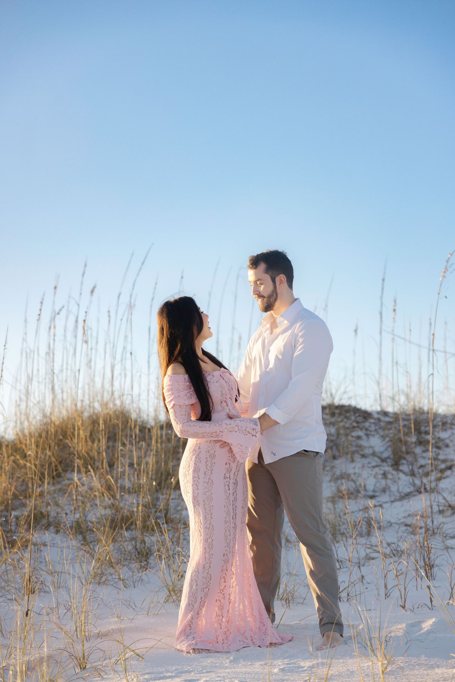 Couple shares romantic moments on a sandy beach with sea oats during sunset engagement photoshoot.