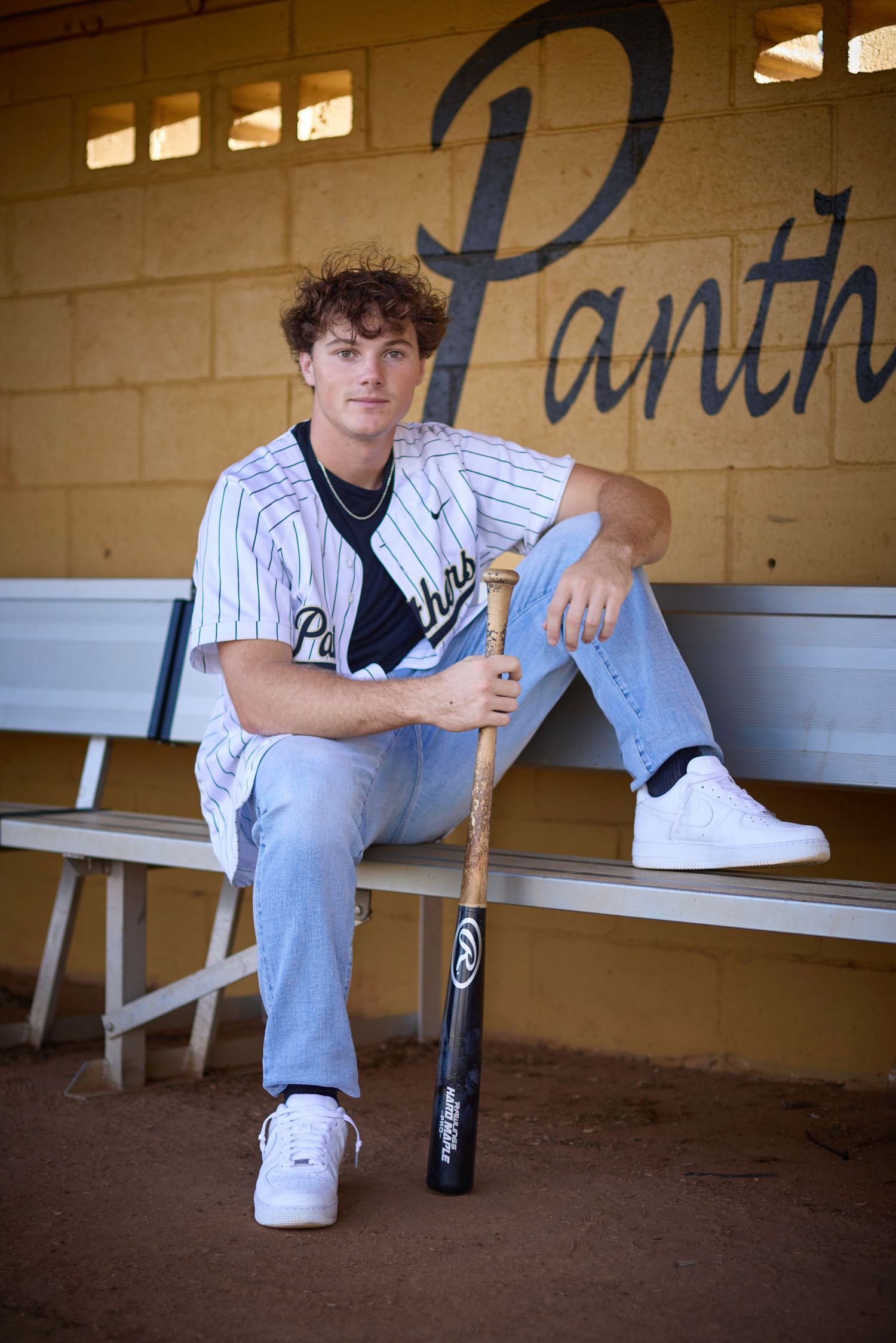 Baseball player in pinstriped uniform sits in dugout holding bat against wall with 'Panth' lettering.