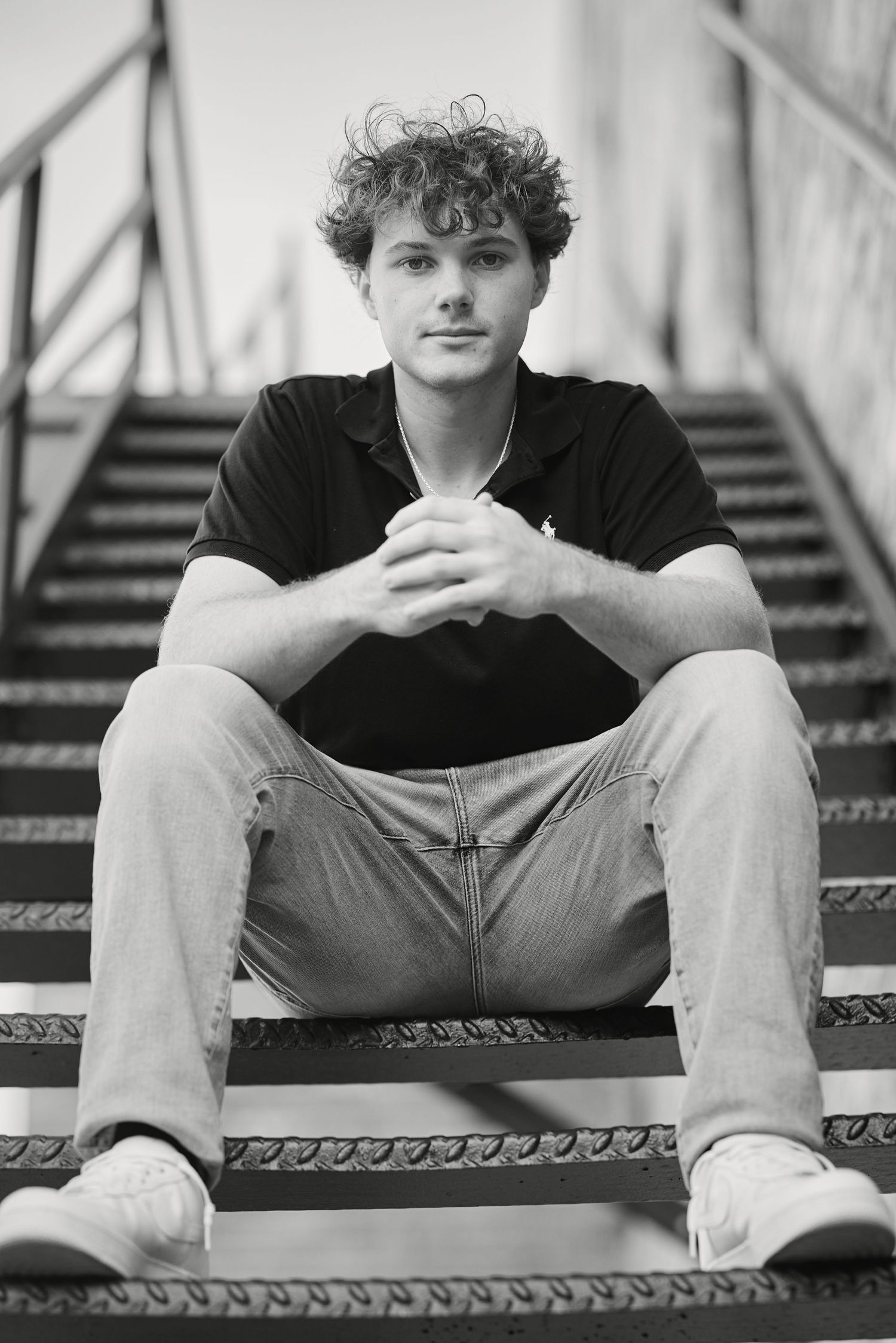 Young person in black shirt sits on metal stairs in black and white portrait.