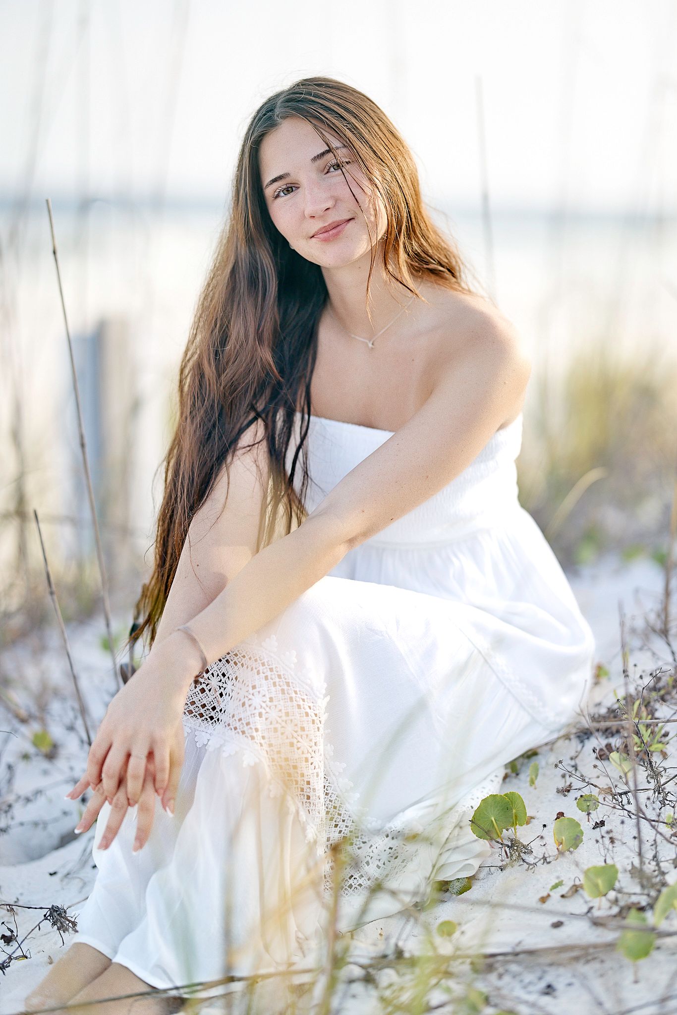 A woman in a white dress sits in soft beach grass during golden hour.
