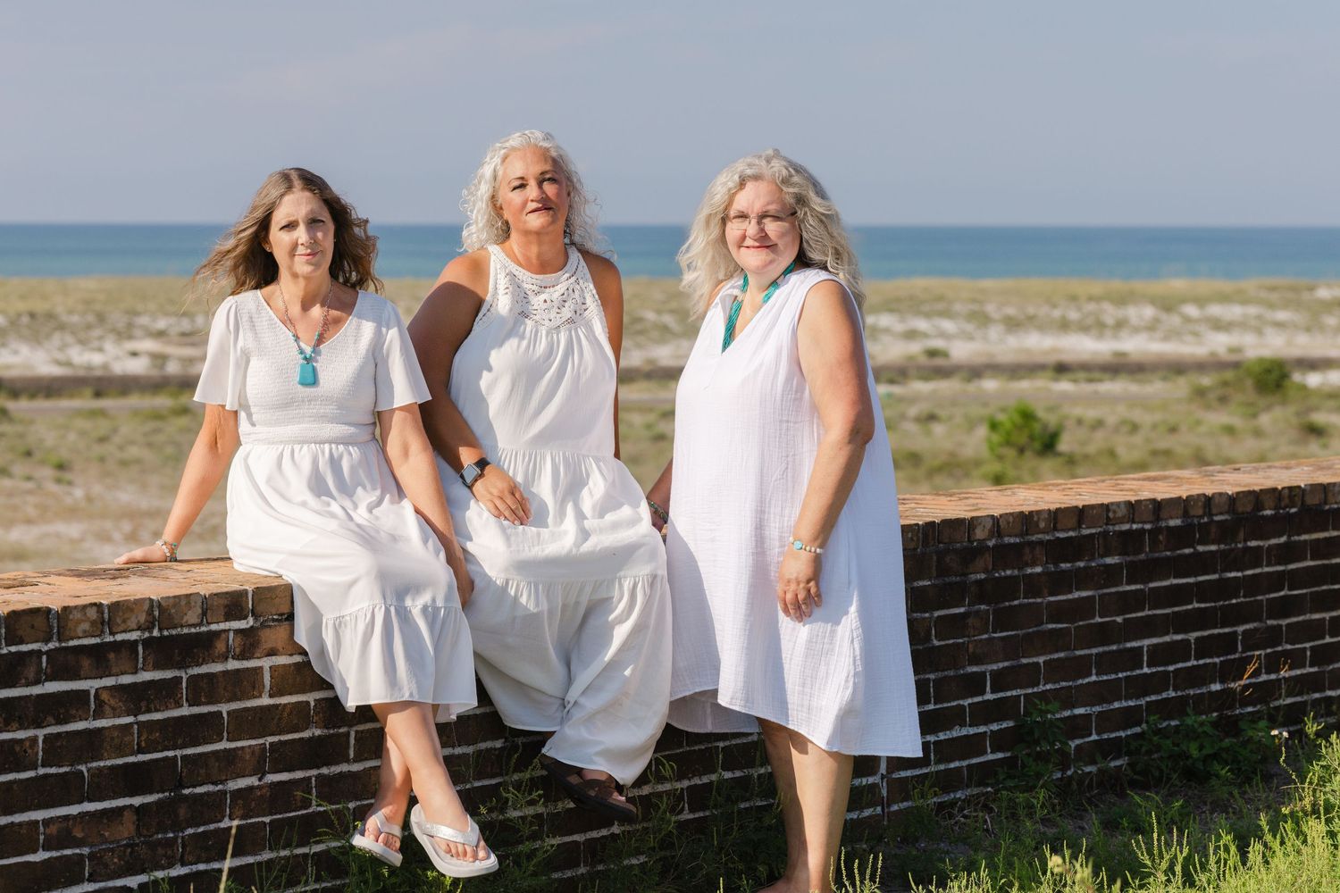 Three people in white summer dresses sit together on a brick wall by the beach on a sunny day.