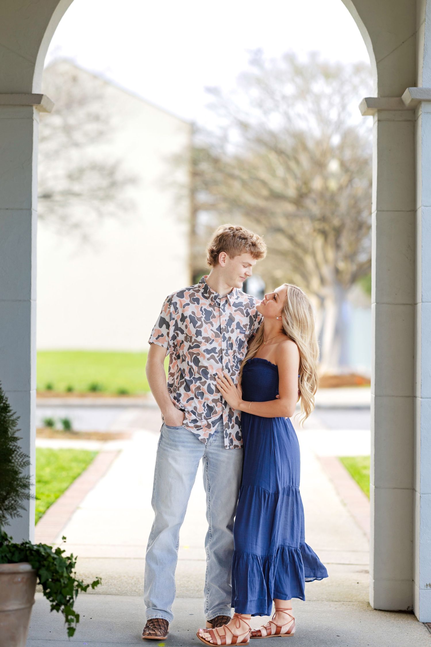 A romantic couple in casual attire share a tender moment under an arched walkway on a spring day.