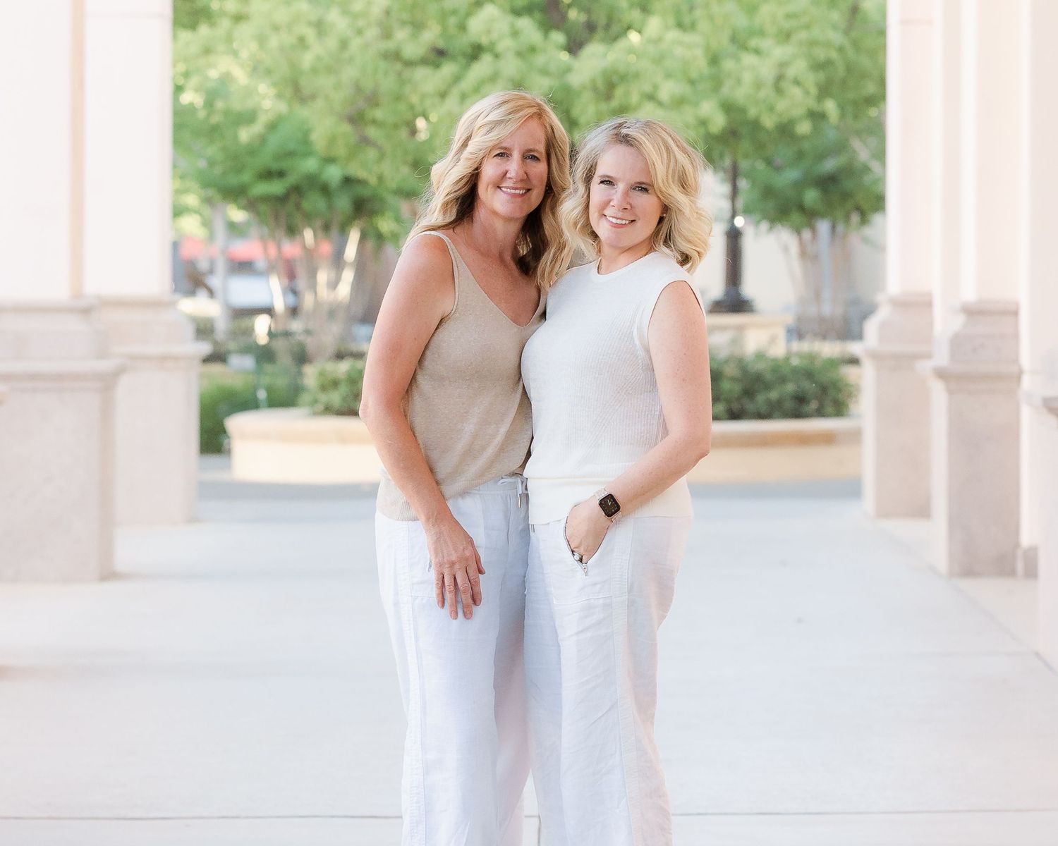 Two smiling women posing together in white and beige outfits for an outdoor portrait.