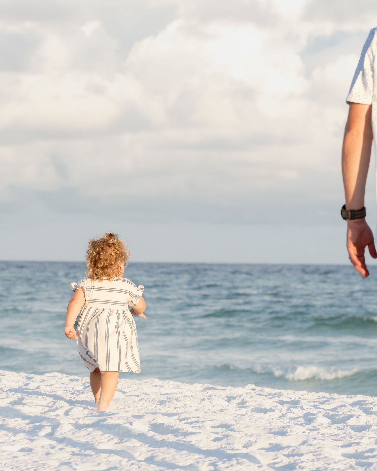 A young curly-haired toddler in a striped dress walks toward ocean waves on a white sandy beach with an adult nearby.