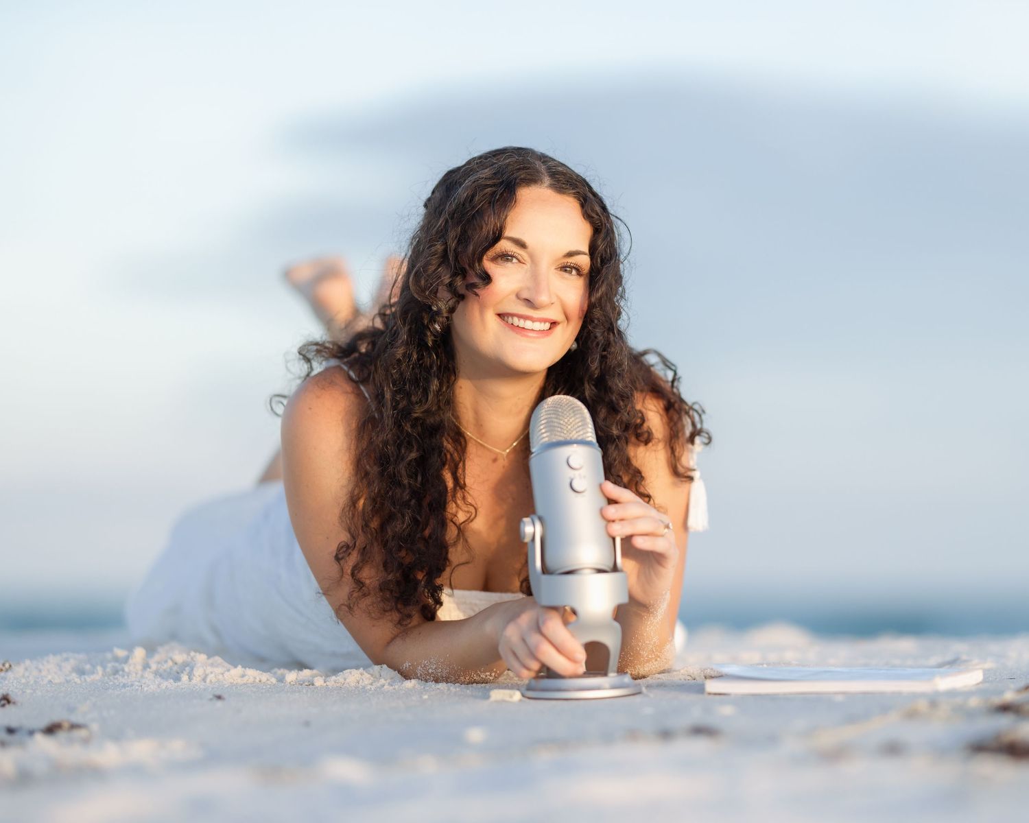A smiling woman with curly hair lies on a sandy beach holding a blue condenser microphone in natural light.