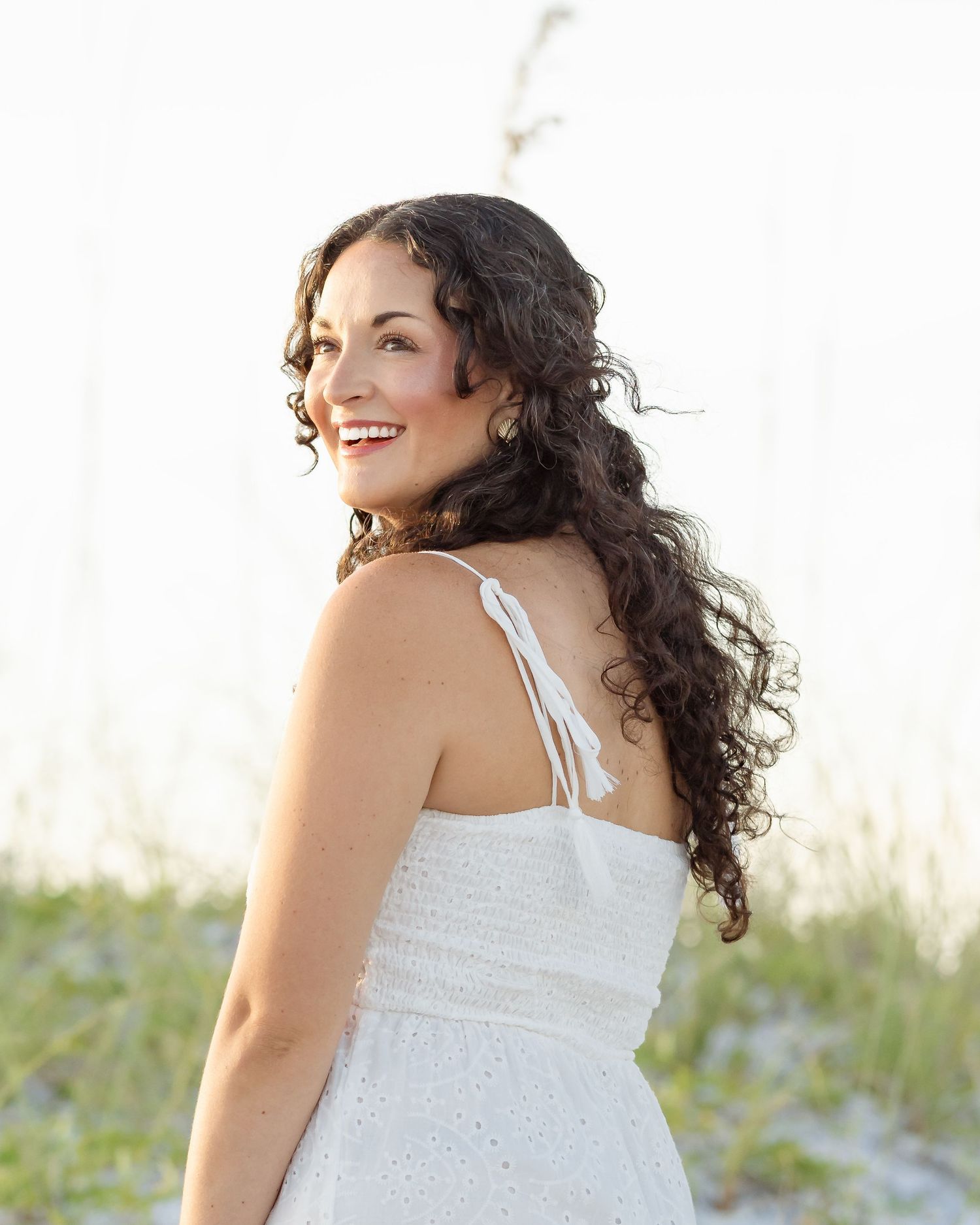 Young woman with curly dark hair in white sundress smiling over shoulder on sandy beach.