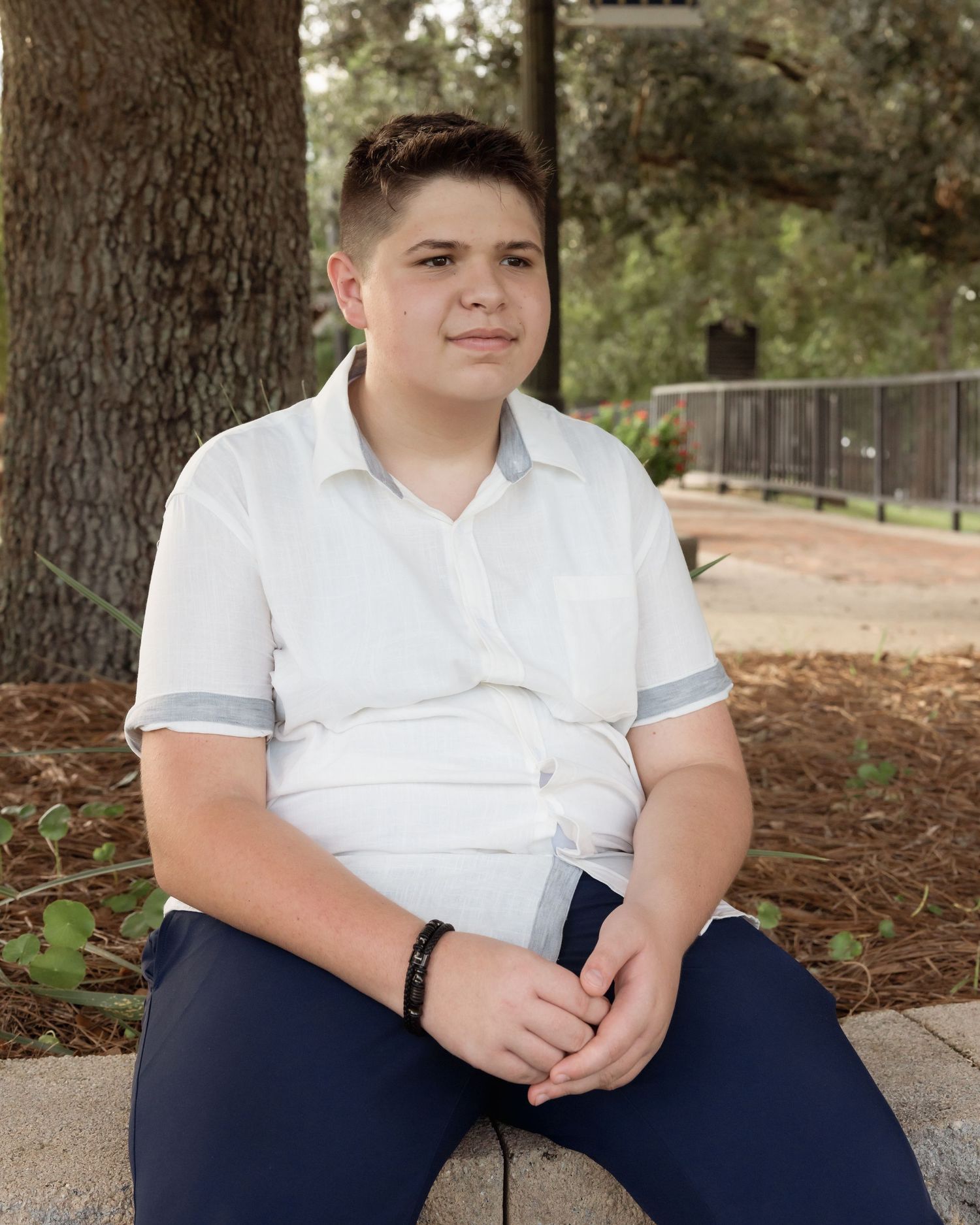 Teen boy in white polo shirt sits on stone bench outdoors, smiling calmly near trees.