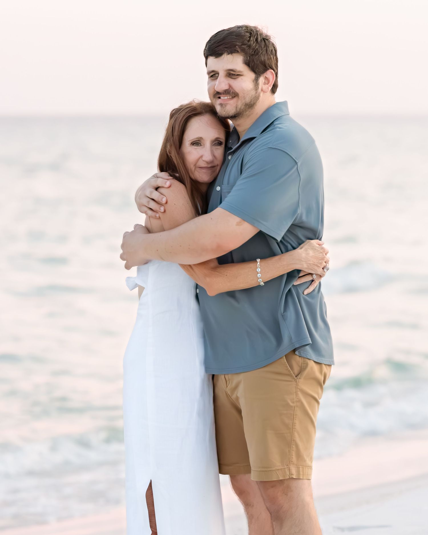 A smiling couple embraces warmly on a beach at sunset, with calm ocean waters in the background.