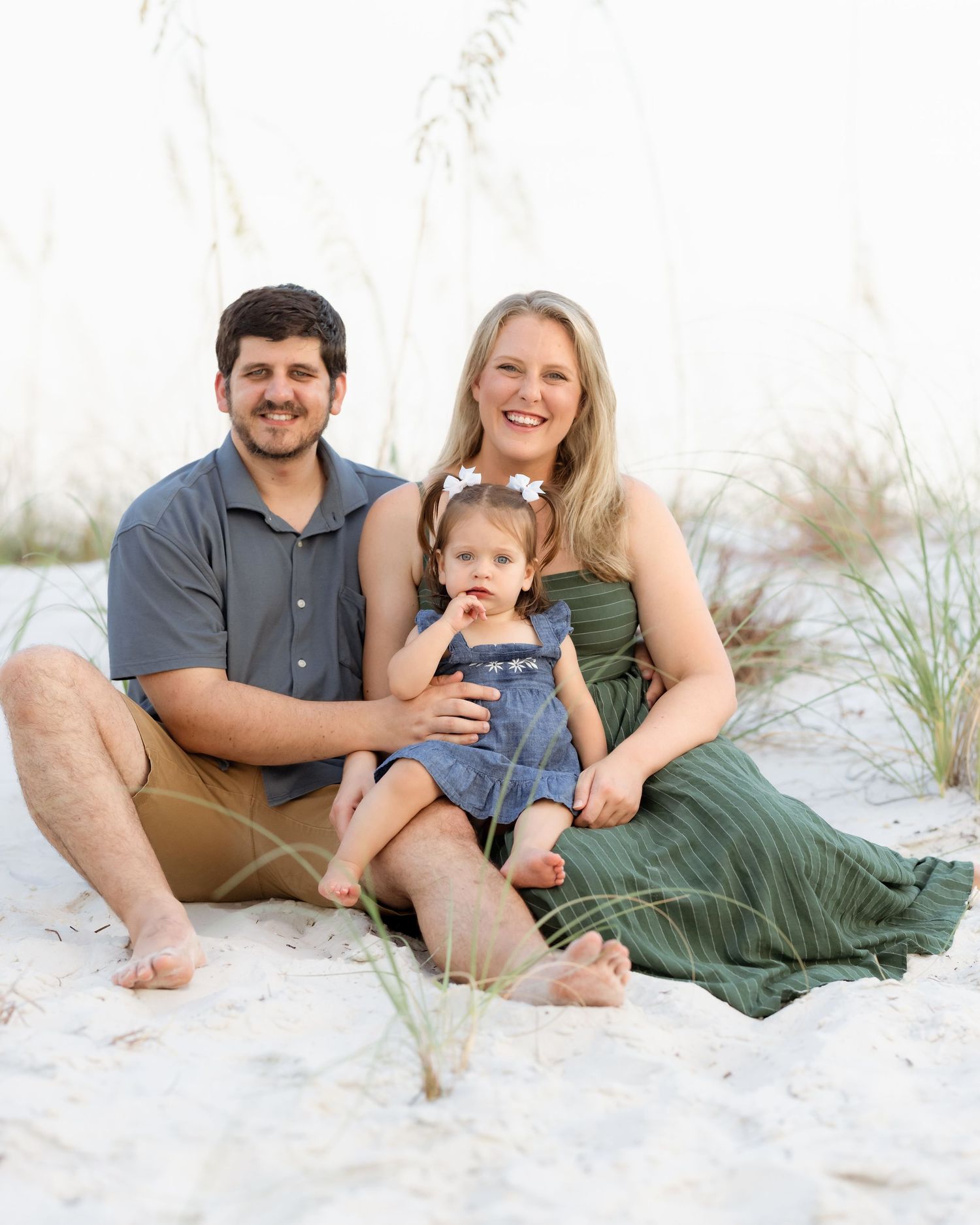 Family of three smiling together on white sand beach, parents sitting with toddler in denim overalls near sea oats.