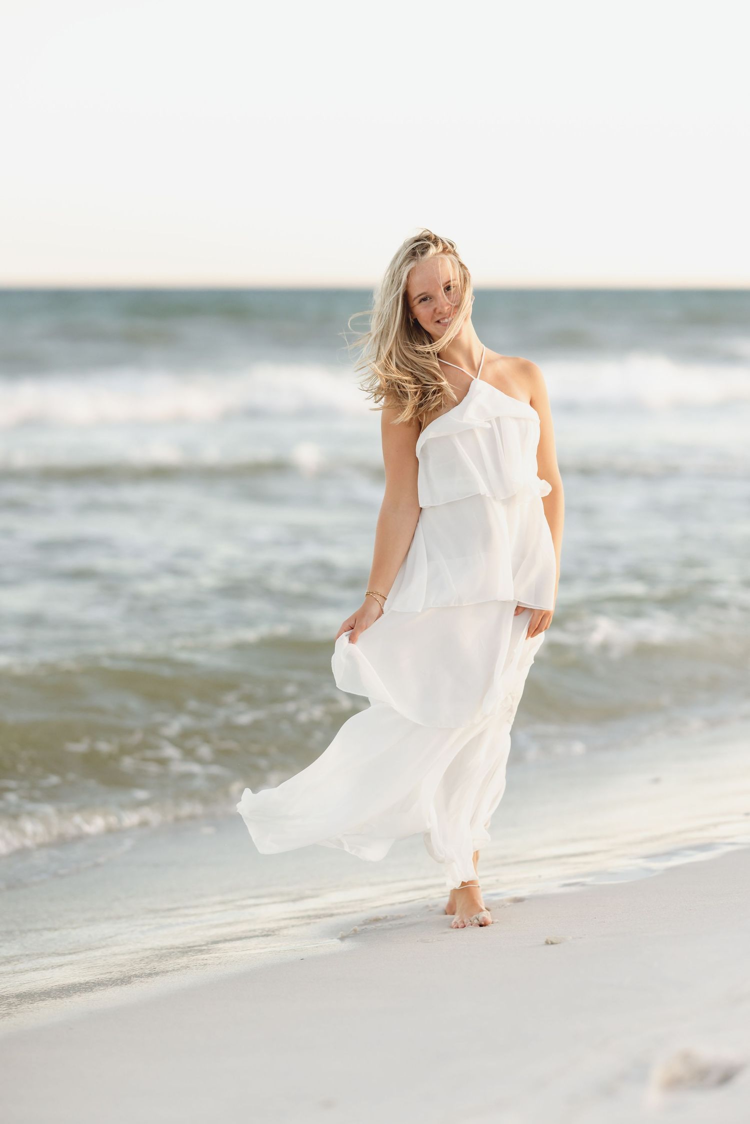A smiling blonde woman in a flowing white maxi dress walks barefoot along a sandy beach with ocean waves behind her.