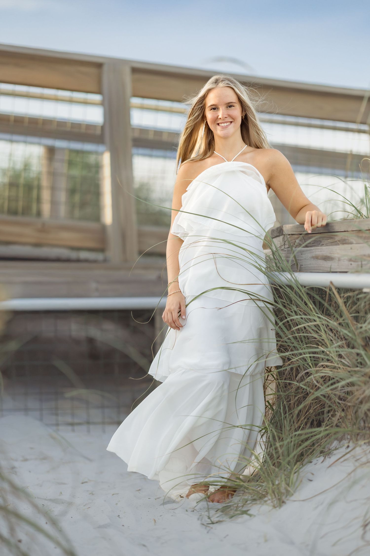 Young woman in white dress smiling on sandy beach boardwalk with sea grass.