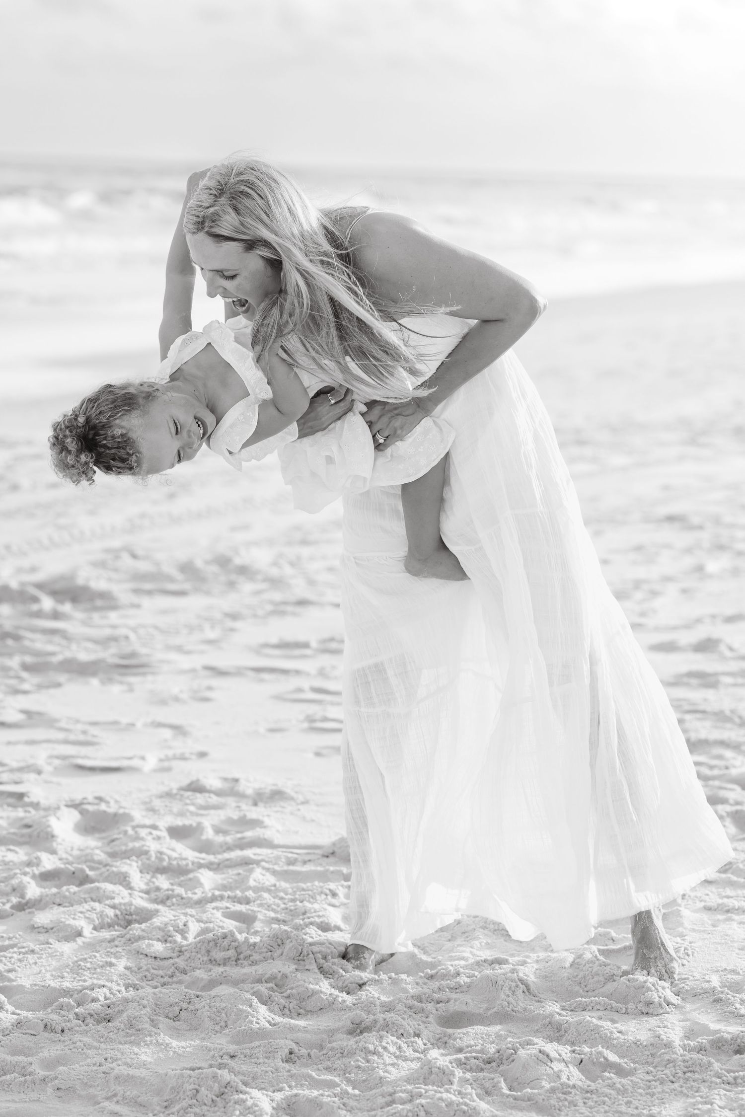 Romantic black and white photo of couple dancing and embracing on a sandy beach in flowing white dress.
