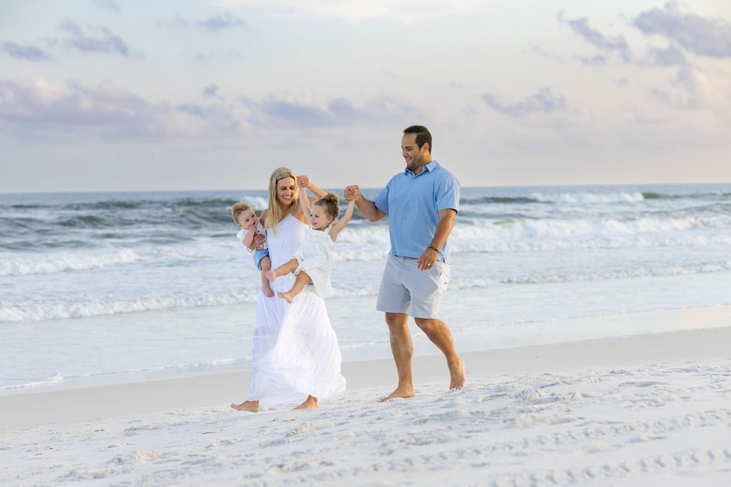 Family of four enjoying a sunny beach walk, parents swinging children on white sandy shore with ocean waves in background.