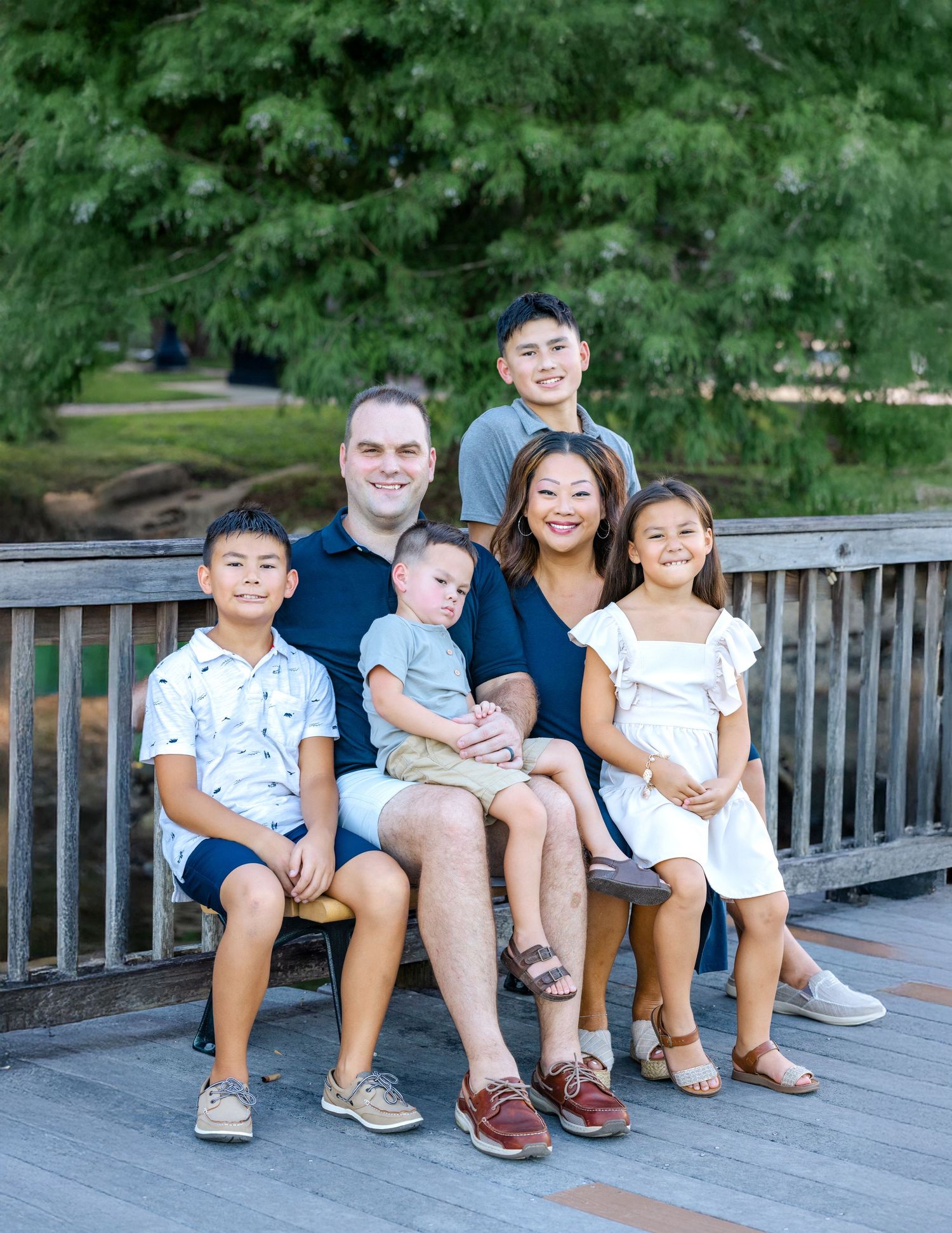 Happy family of six posing together on a wooden bridge outdoors, surrounded by lush green trees.