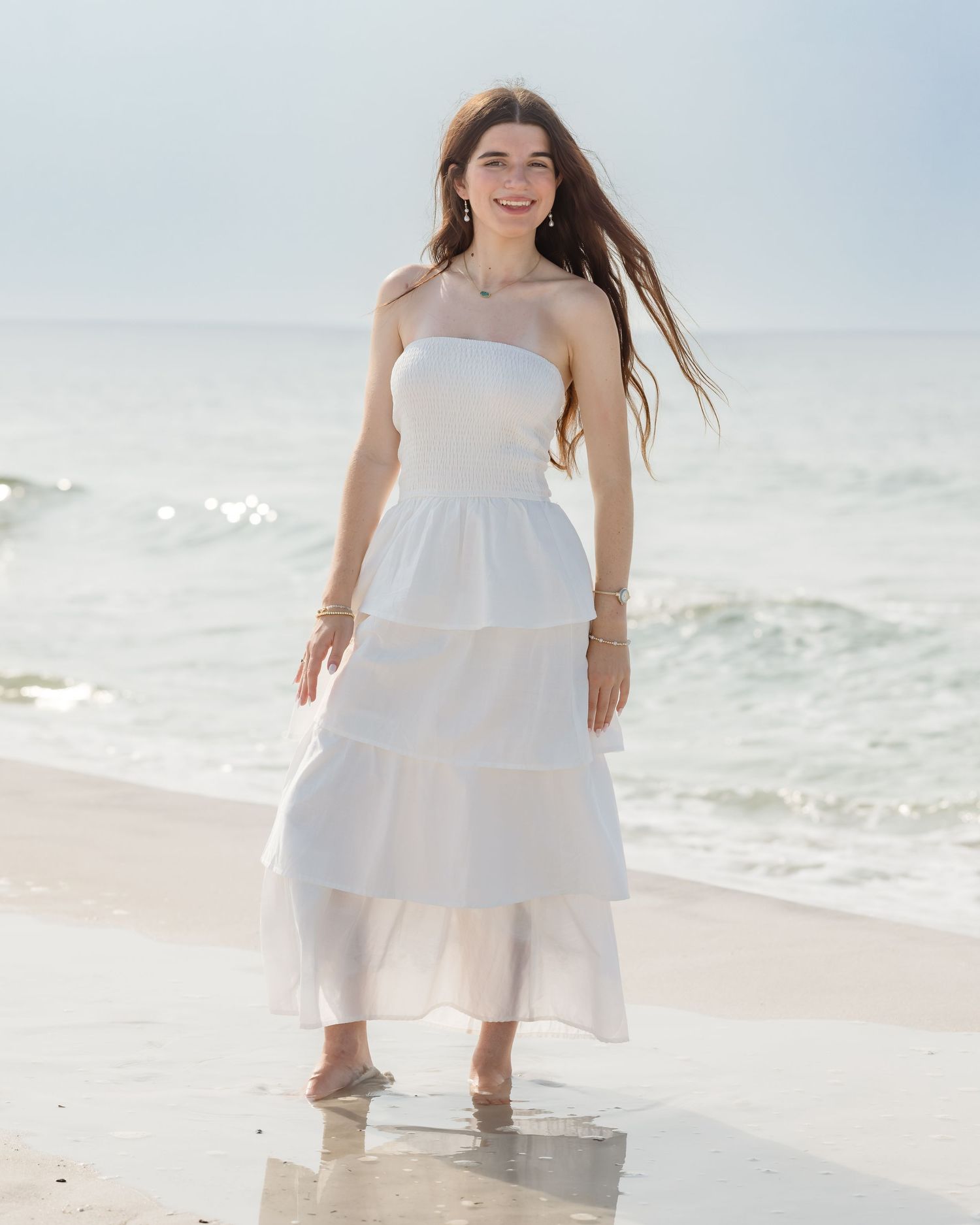 Young woman in white strapless tiered maxi dress standing barefoot on sandy beach with ocean waves behind her.