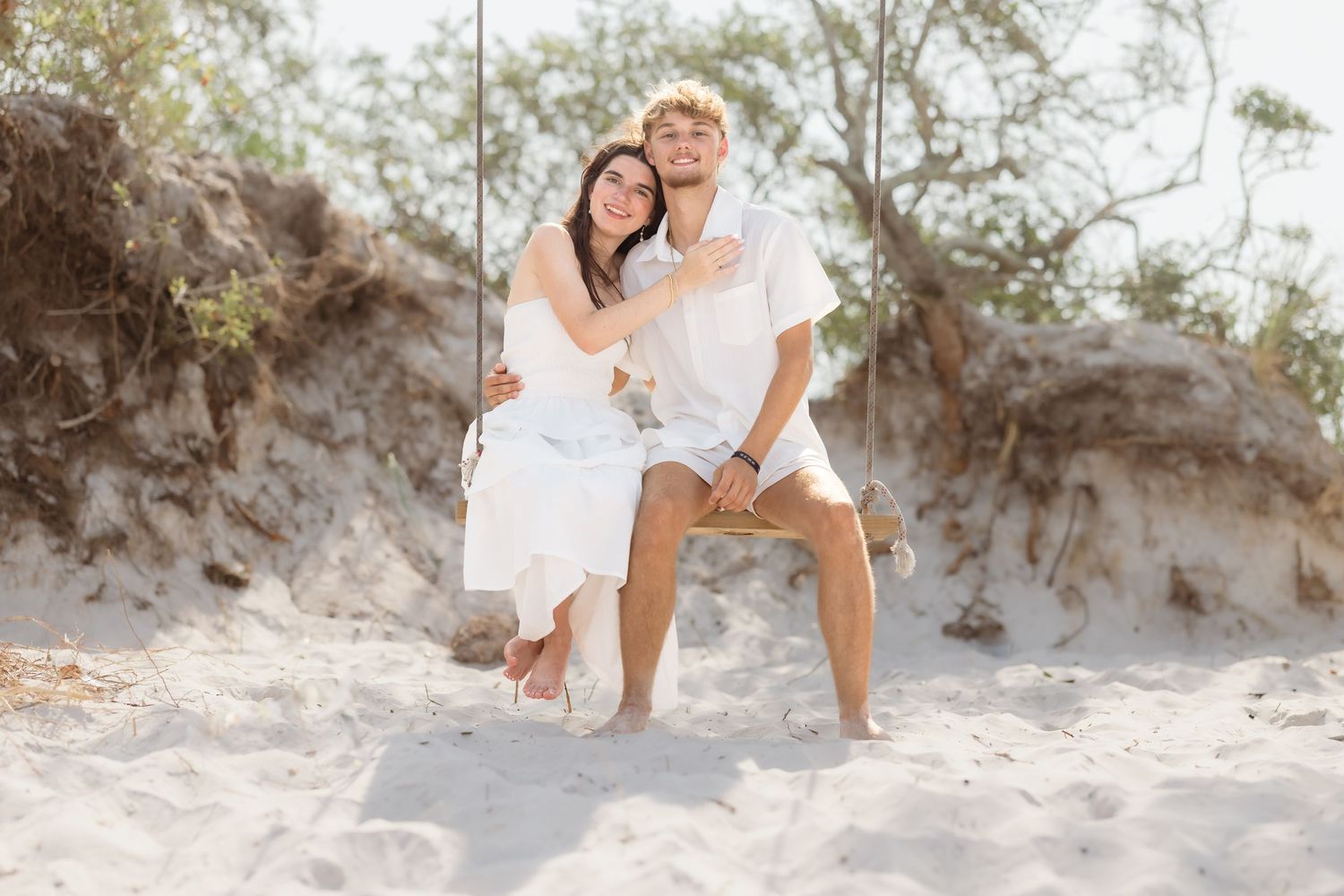 Couple in white outfits sitting on a beach swing surrounded by sand dunes and trees on a sunny day.