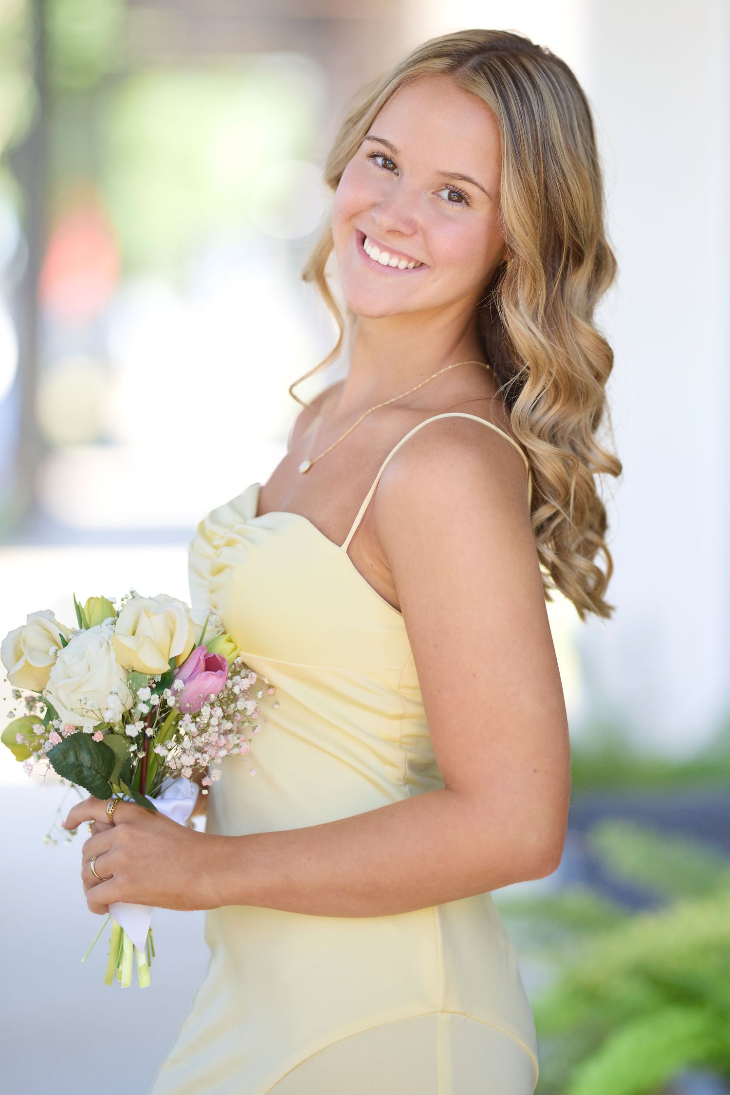 Person in yellow dress holding white and pink floral bouquet poses with curled hair in outdoor setting.