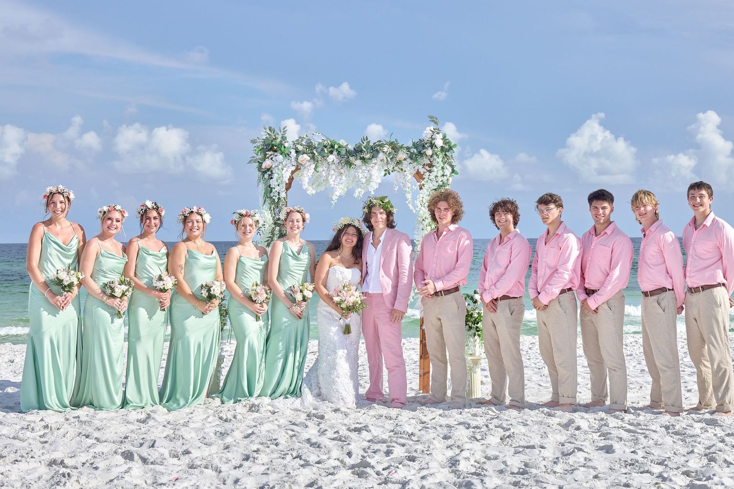 A wedding party wearing pastel pink and mint green dresses and light colored suits poses on a sandy beach.