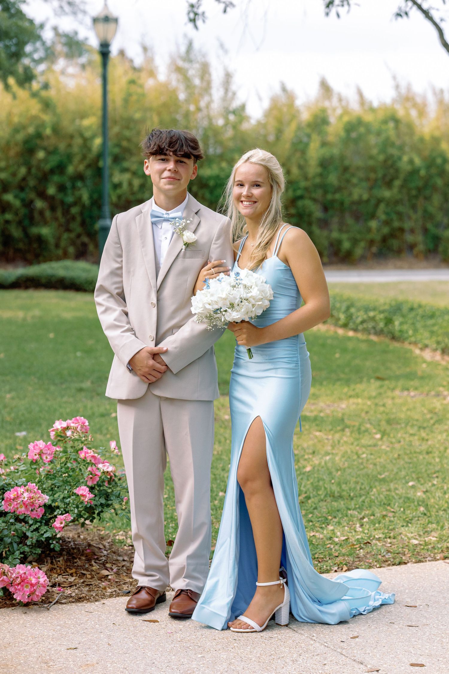 A couple poses outdoors at a formal event, with her in a light blue gown and him in a beige suit next to pink flowers.