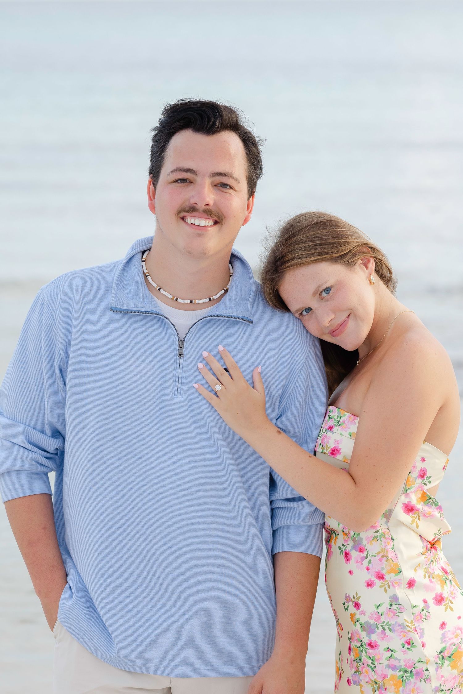 Happy couple in floral dress and blue polo embracing by the ocean.