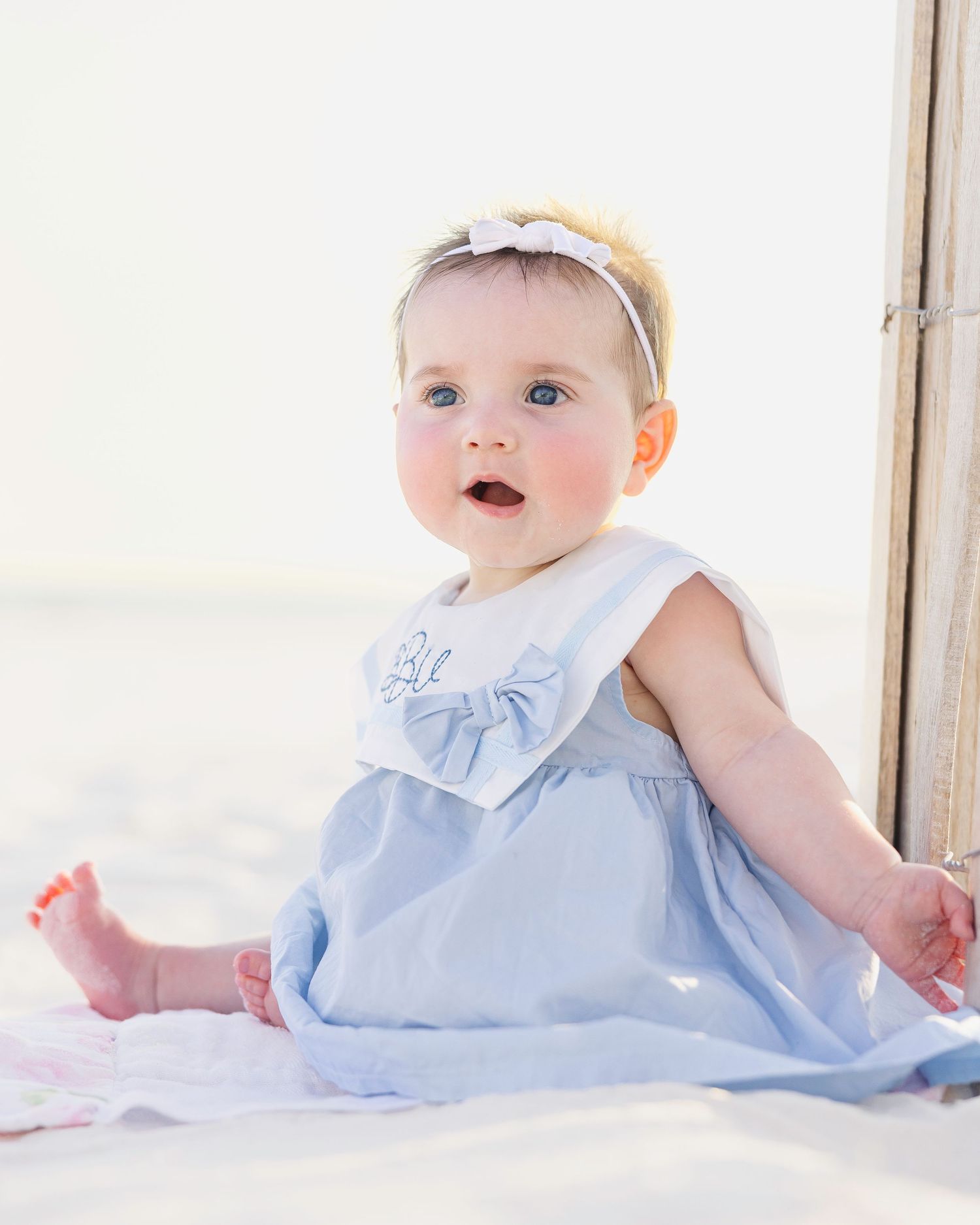 Baby in pale blue dress sitting on soft white blanket in bright natural light for professional photo.
