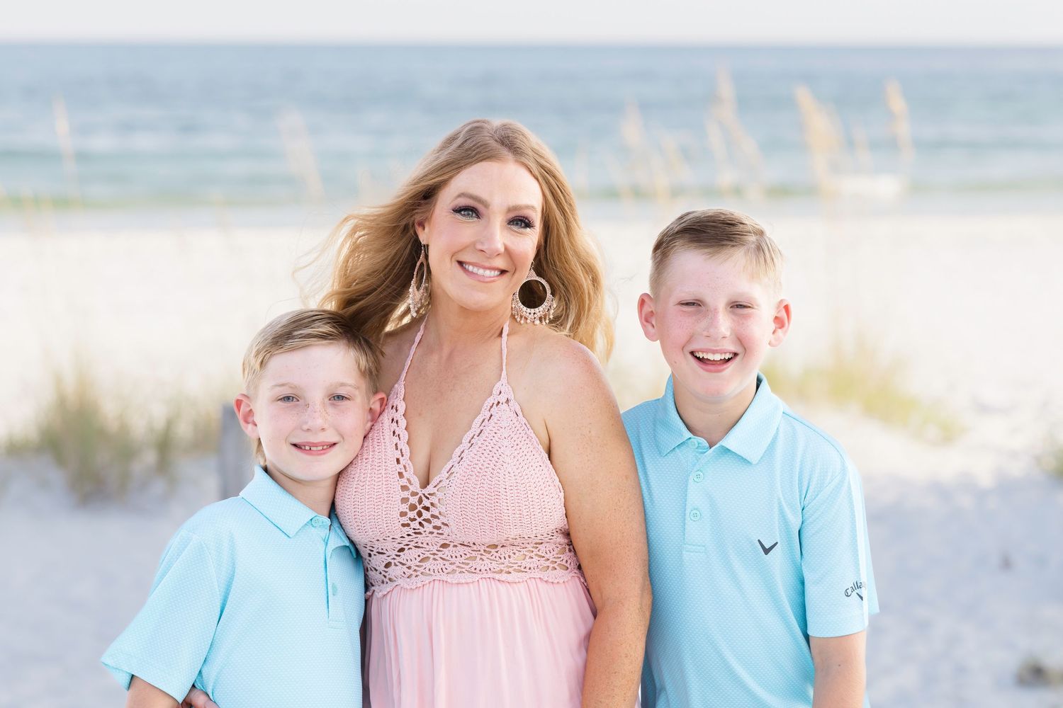 Family portrait session on a sunny beach with people wearing coordinated pink and turquoise outfits.