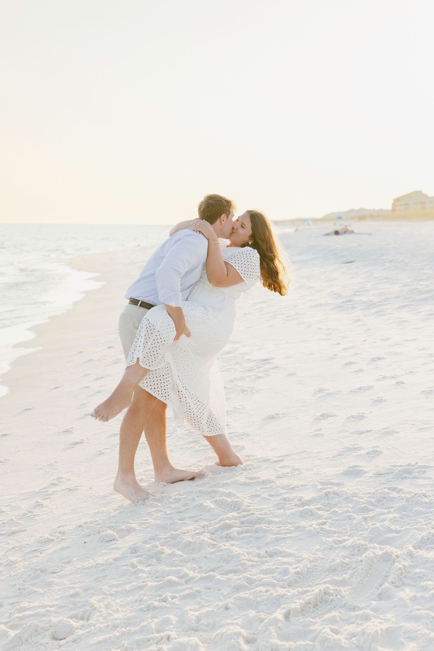 A romantic couple in white outfits dances and embraces on a sunny beach during sunset.