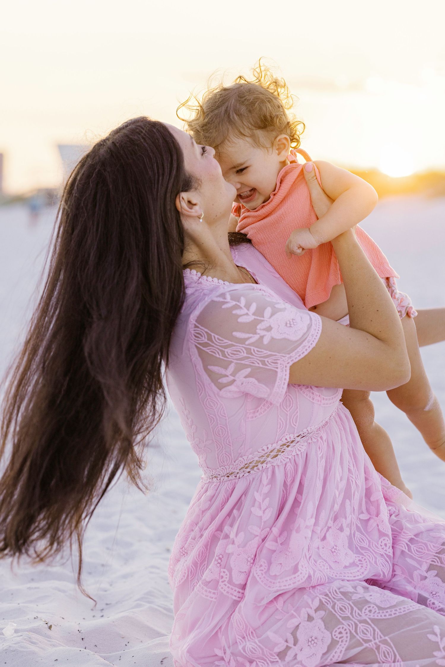 Mother and baby in pink dress share tender moments on the beach at sunset.